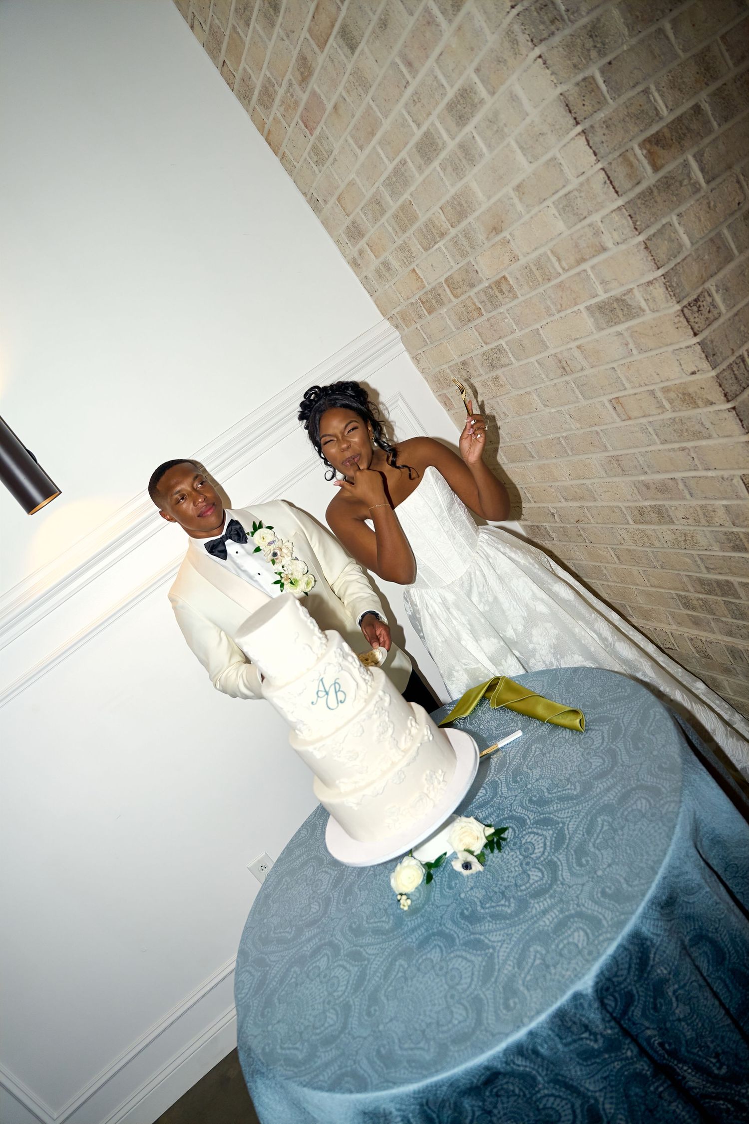 Bride and groom pose beside elegant four-tier white wedding cake on blue tablecloth against brick wall.