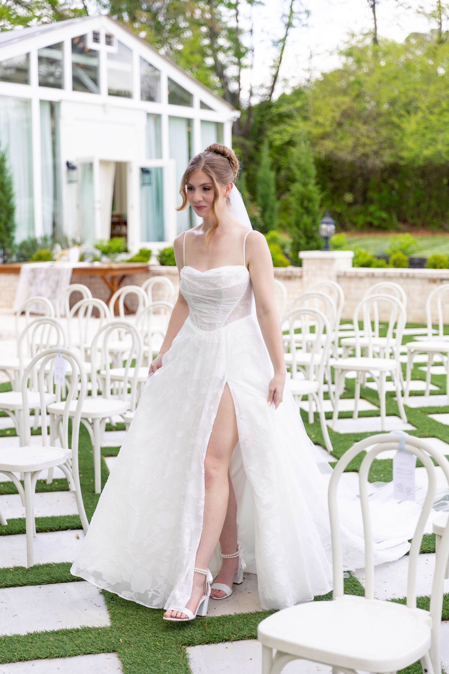 Elegant bride in white ball gown with slit and veil standing among white chairs at outdoor wedding venue.