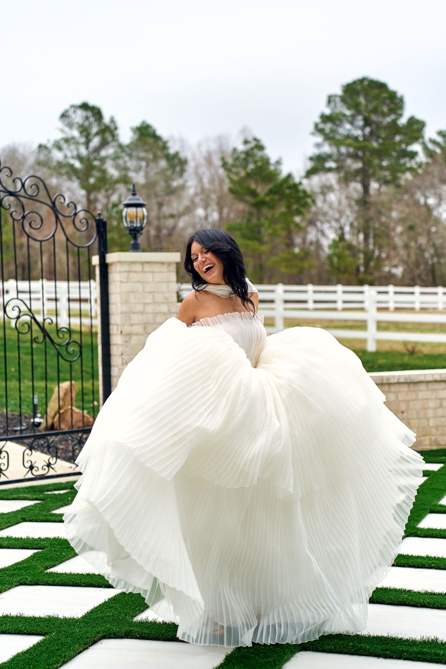 A white pleated wedding gown swirls dramatically on a green lawn with decorative gate and trees in the background.