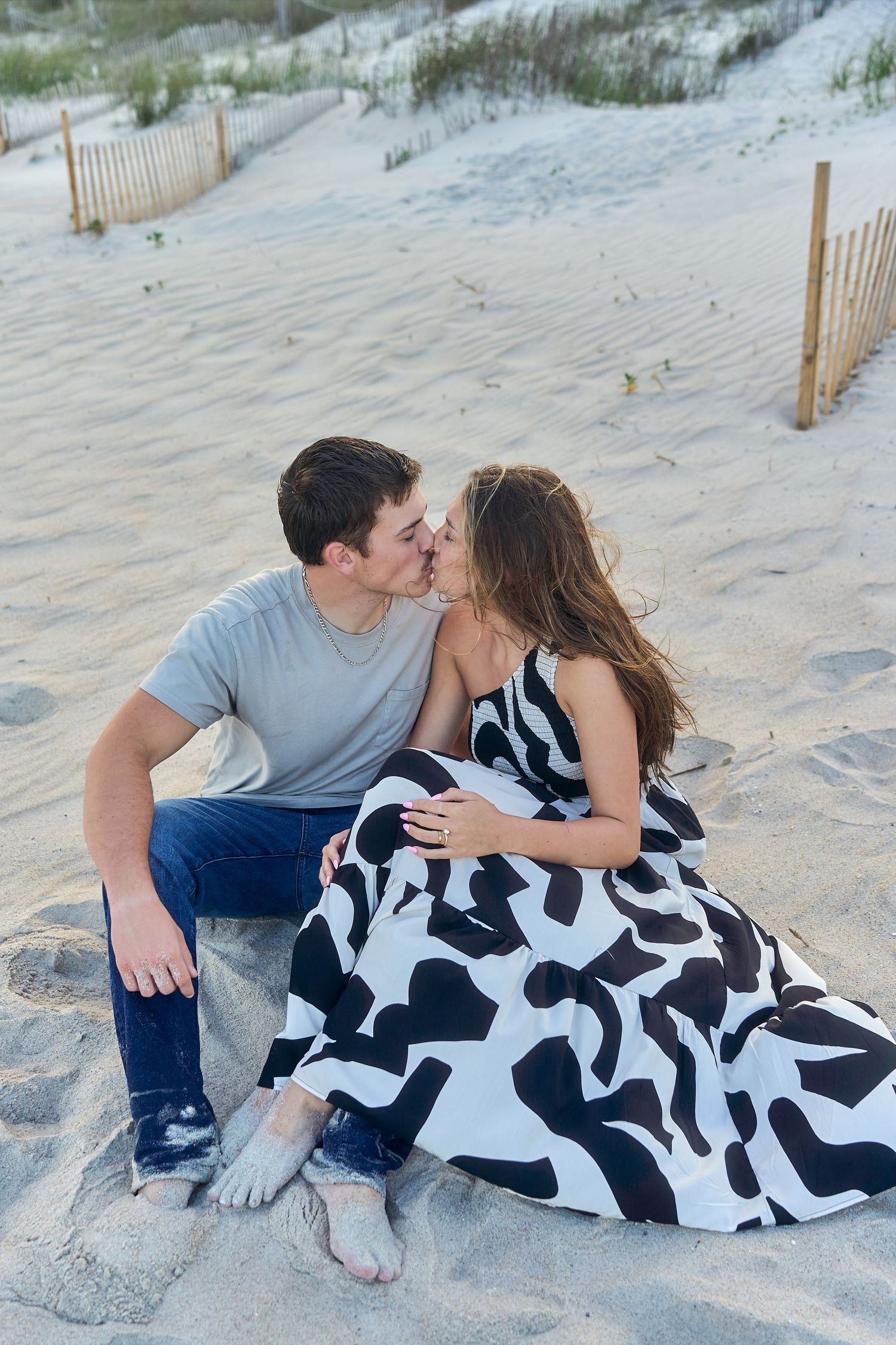 A couple shares a romantic moment sitting on sandy beach dunes with wooden fences in the background.