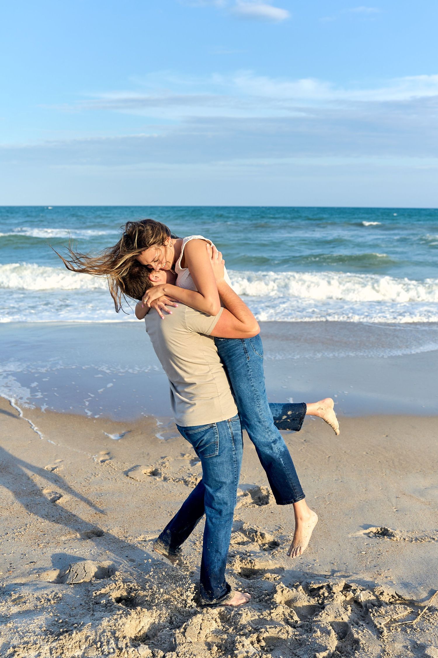 Romantic couple embracing and kissing on sandy beach with ocean waves and blue sky in background.