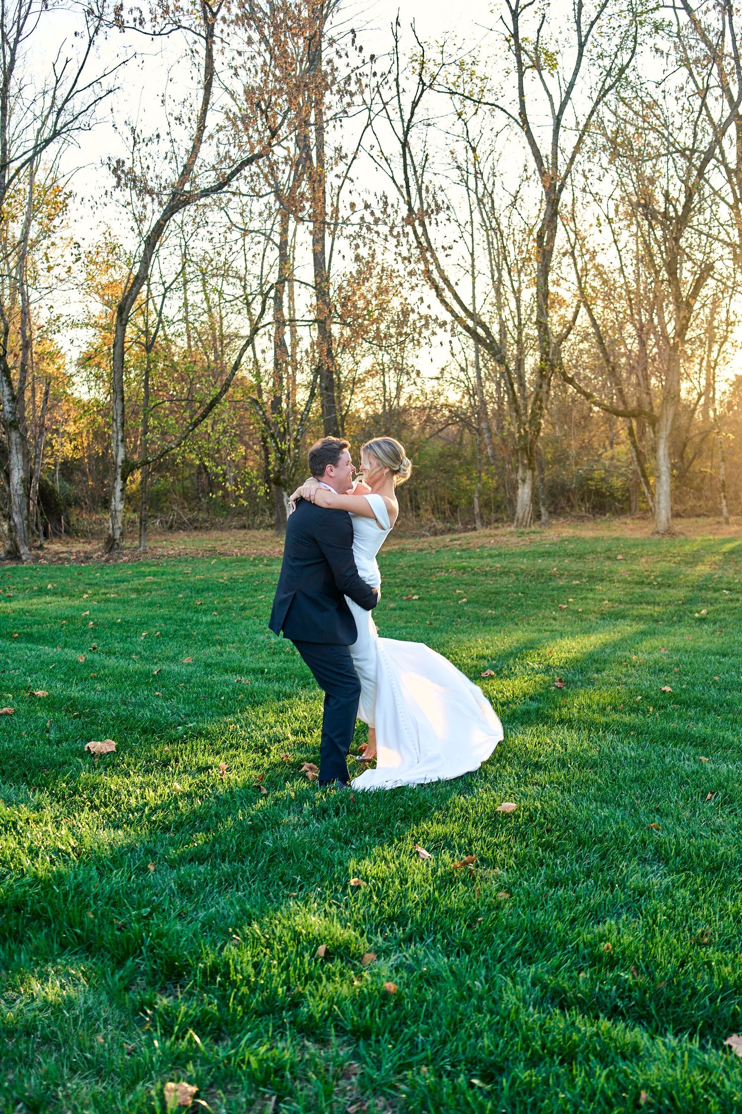 A couple shares a romantic embrace on a lush green lawn at sunset with bare trees in the background.