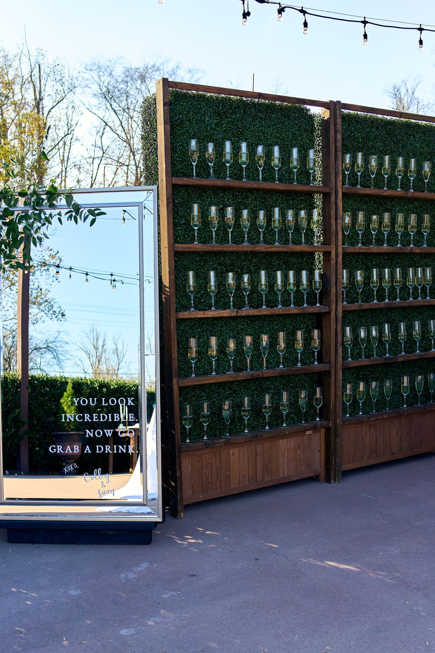 A wooden display shelf holds rows of green glass bottles with string lights overhead and a sign inviting people to grab a drink.
