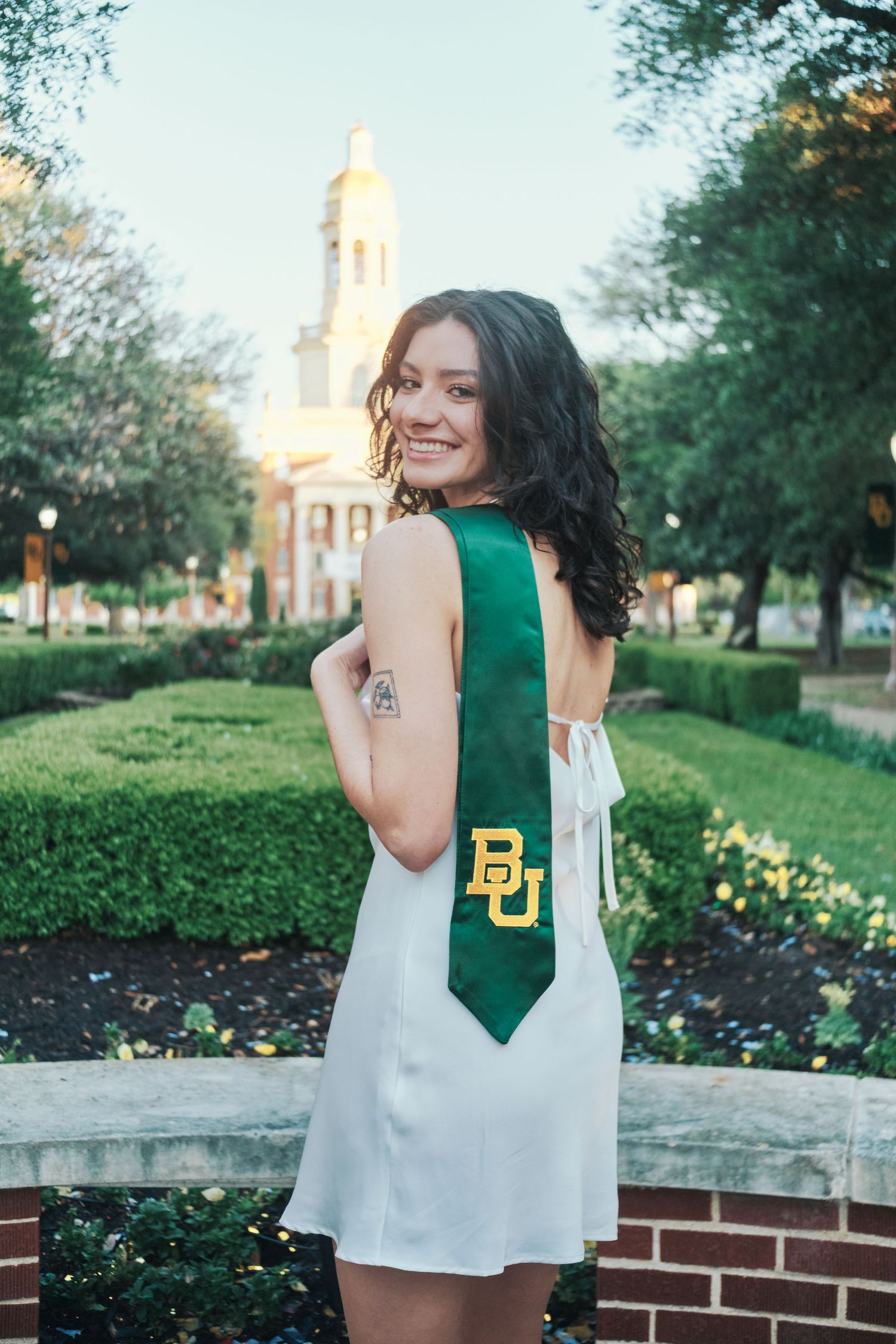 Brunette woman in white dress showing BU green graduation stole near Baylor chapel.