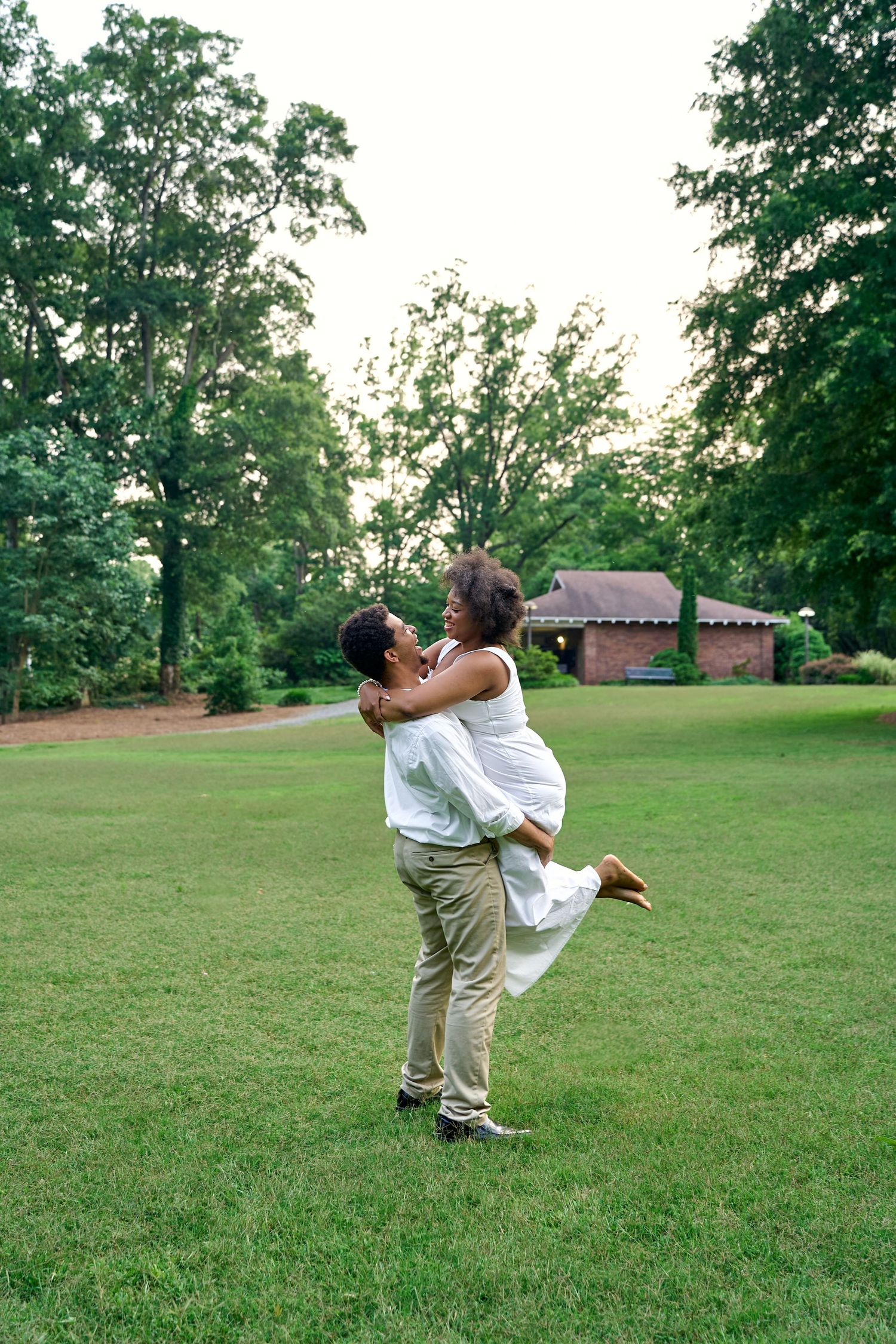 A playful couple dances and spins together on a grassy lawn surrounded by trees on a summer evening.