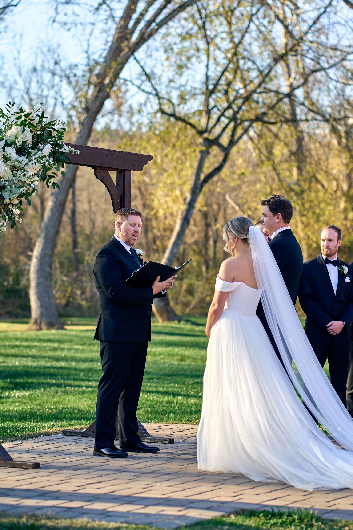 An outdoor wedding ceremony taking place on a wooden platform surrounded by autumn trees during golden hour.