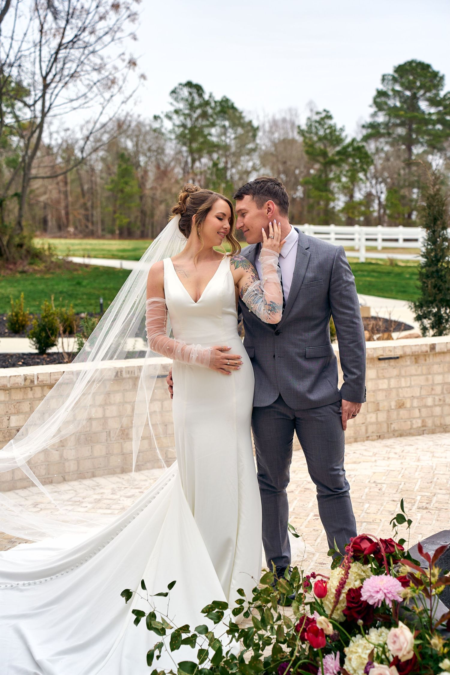 A wedding couple shares an intimate moment on a brick patio with white fence and floral arrangements in the background.