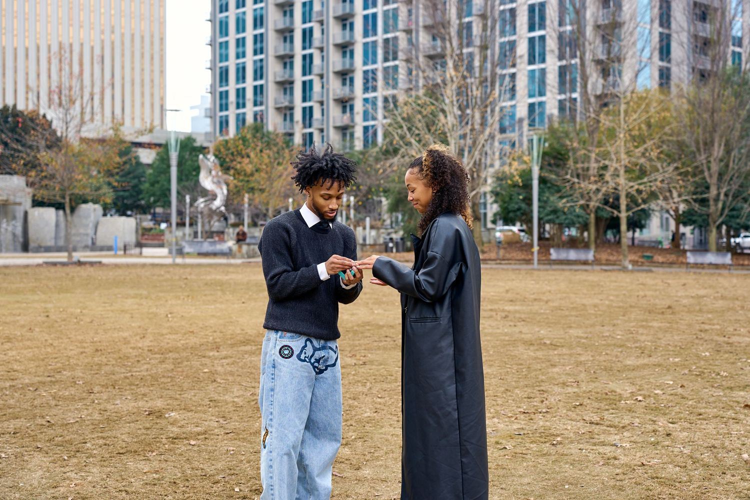 Two people stand together in conversation on a grassy field with tall buildings visible in the background. North Carolina Photographer.