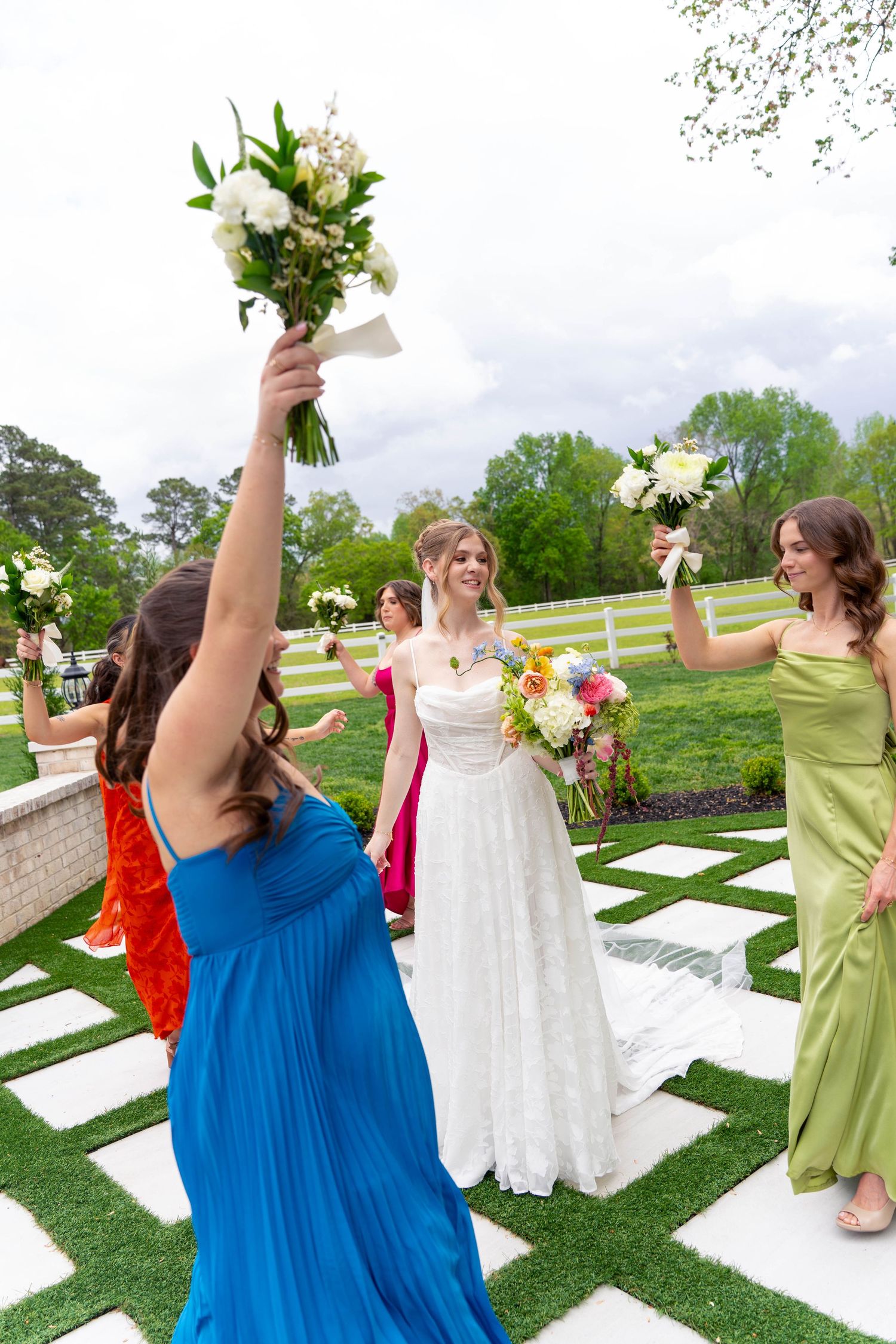 Bride in white lace gown celebrated by colorful bridesmaids raising bouquets at outdoor ceremony.