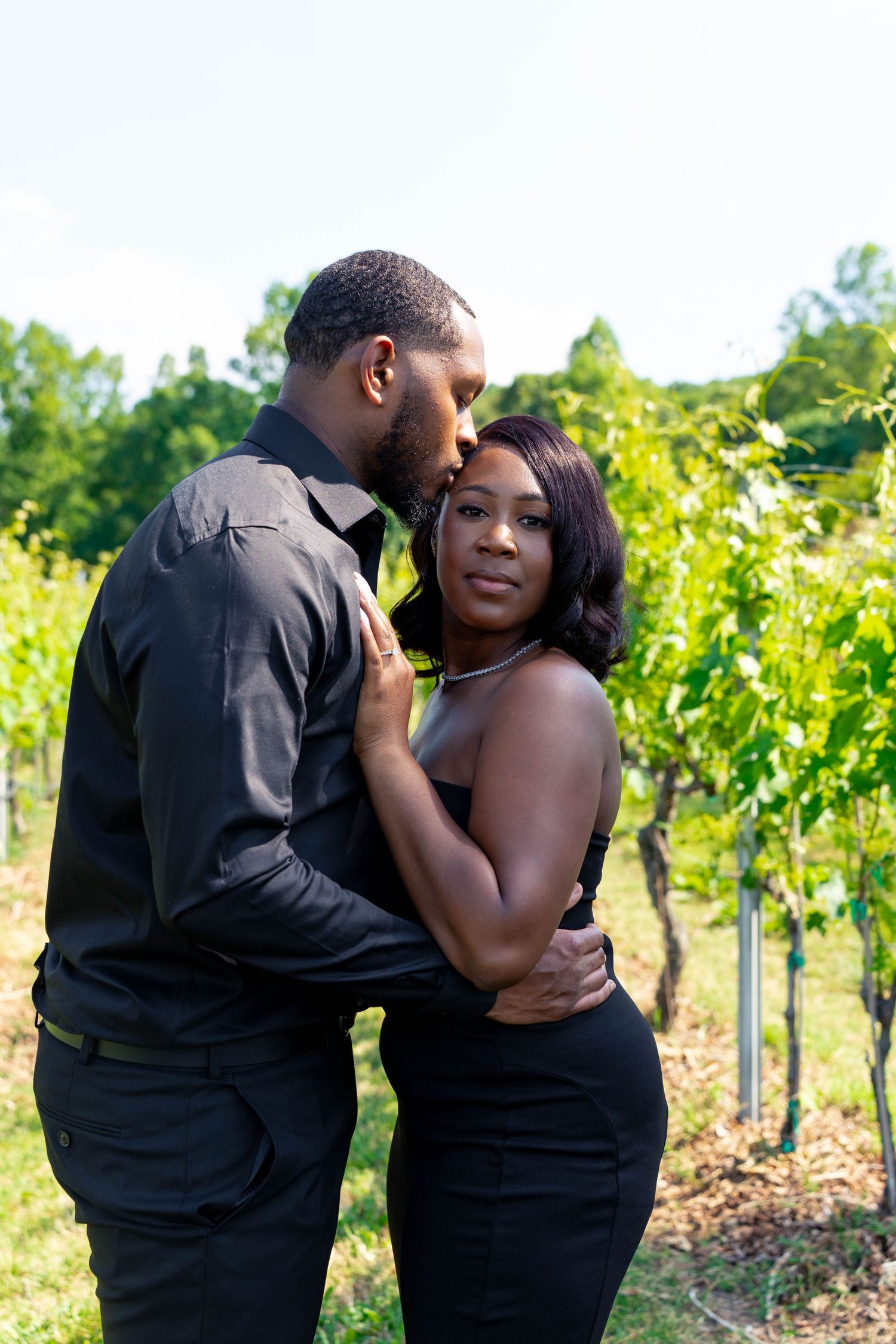 Romantic couple embracing in vineyard, man kissing womans temple while she looks at camera.