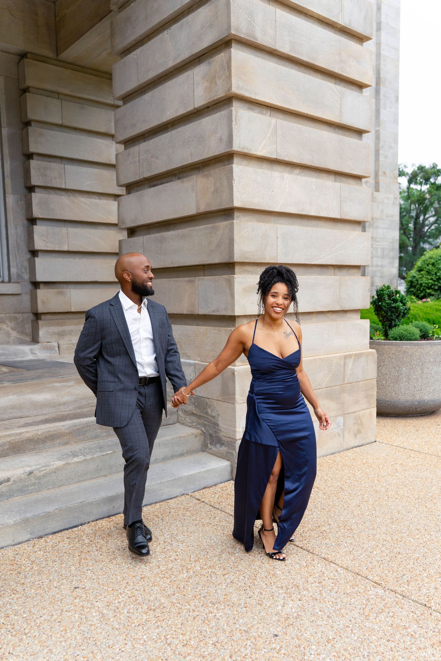 Elegant Black couple holding hands walking past stone columns, woman in navy satin gown, man in gray suit.