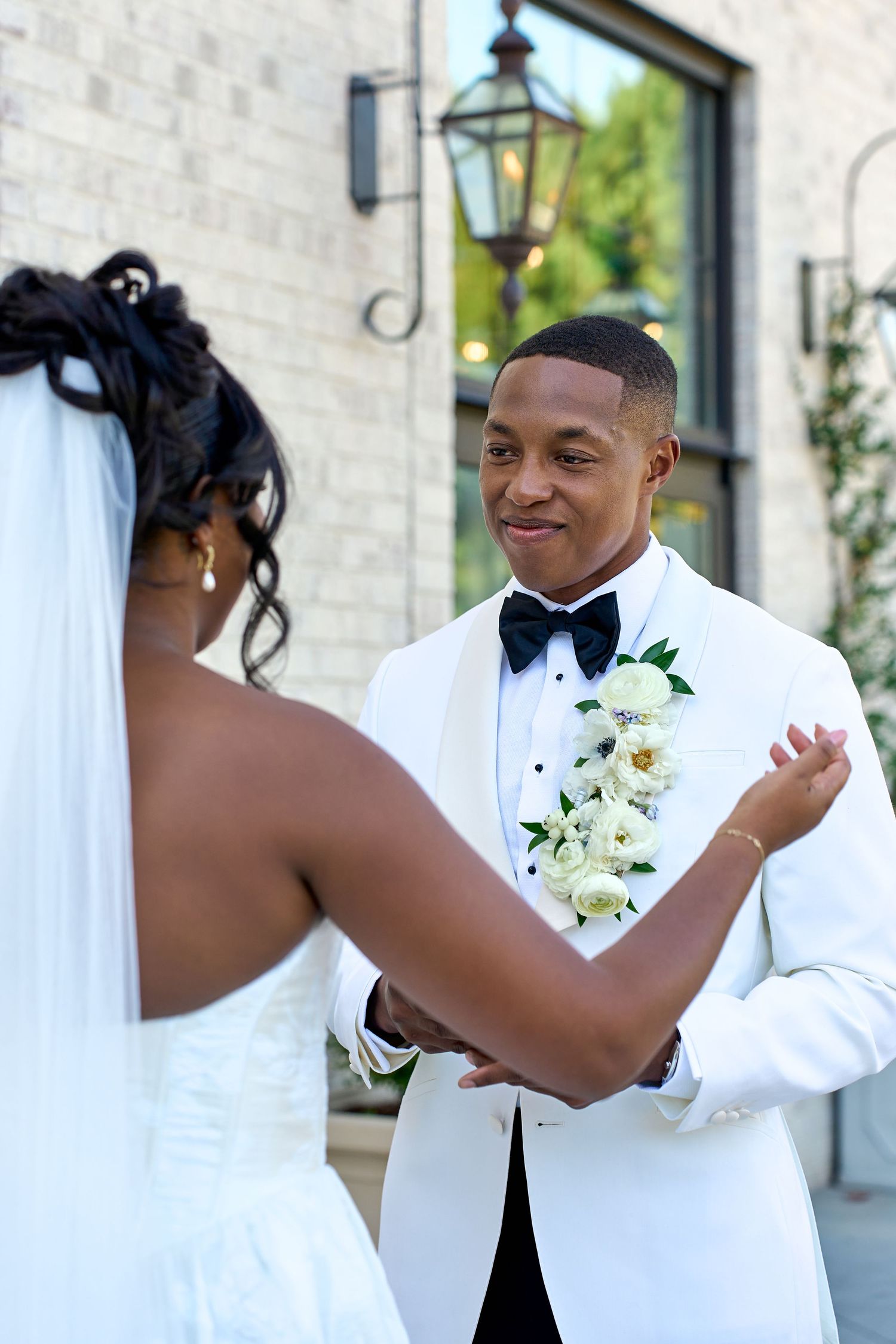 Groom in white tuxedo jacket with floral boutonniere smiles at bride during outdoor first look.