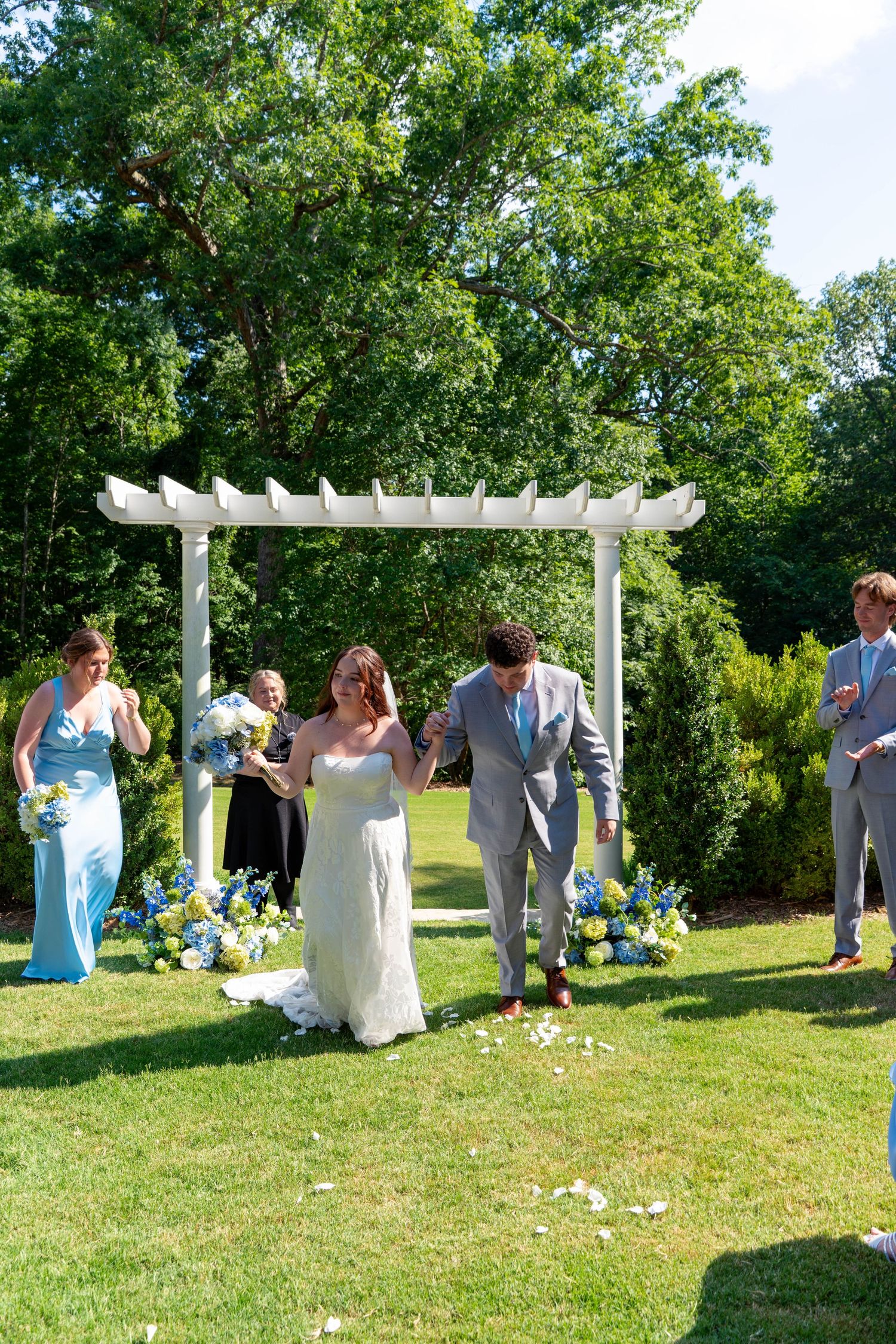 Bride and groom exit ceremony under white pergola arch surrounded by blue and yellow floral arrangements.