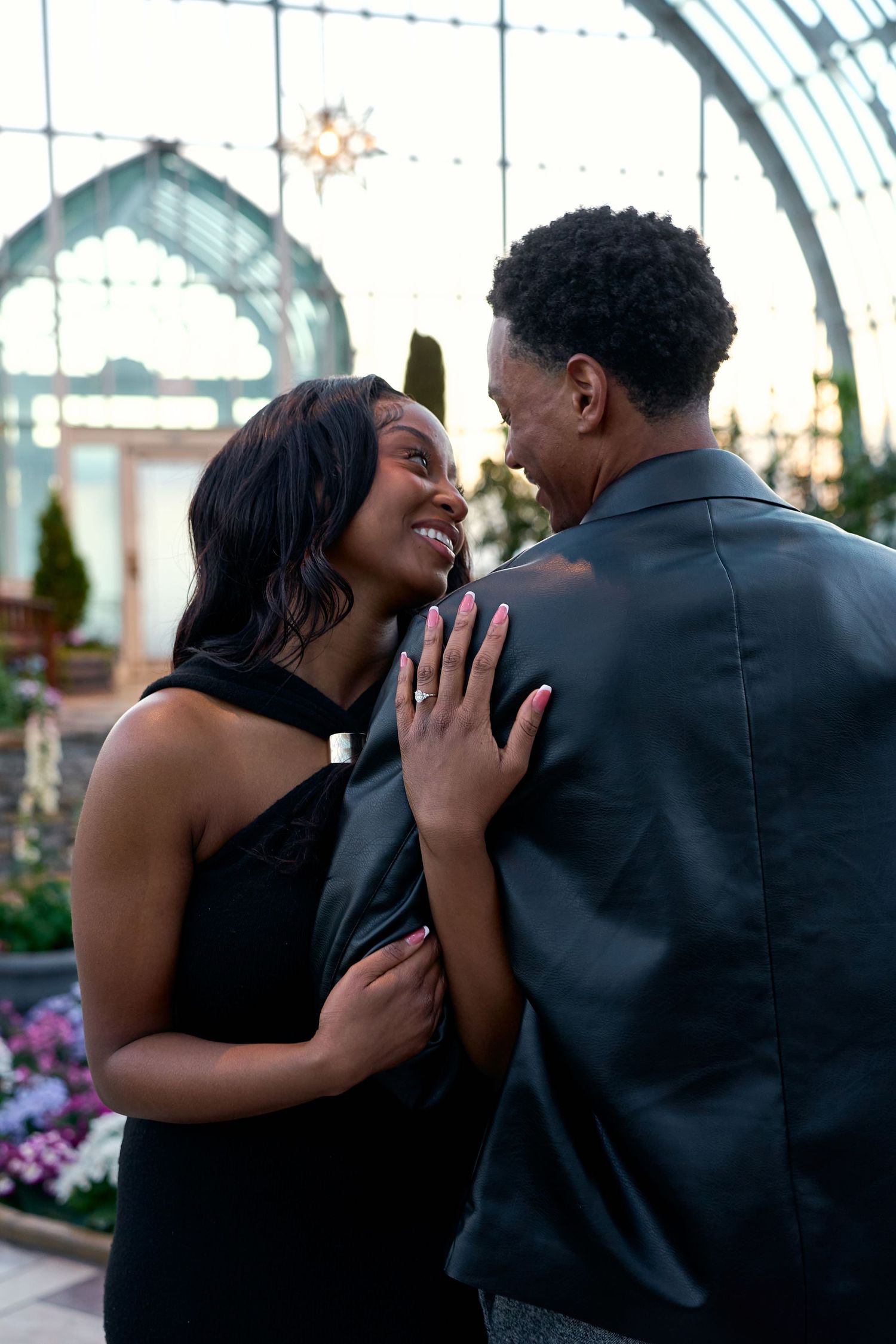 Joyful couple embracing and laughing inside a glass greenhouse with lush green plants behind them.