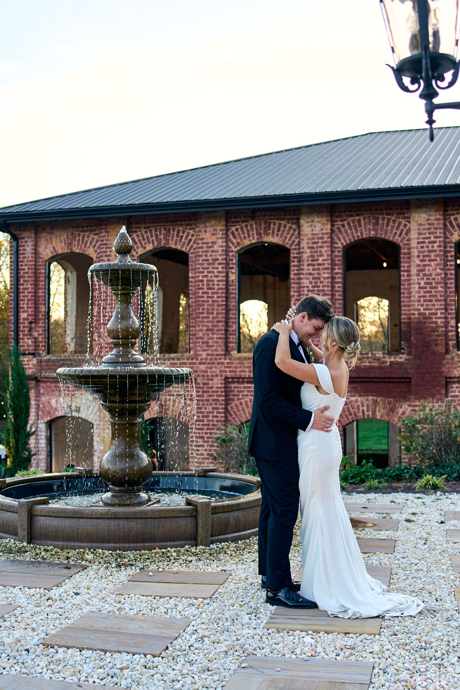 Elegant couple embraces by ornate stone fountain outside rustic brick wedding venue with arched portico.