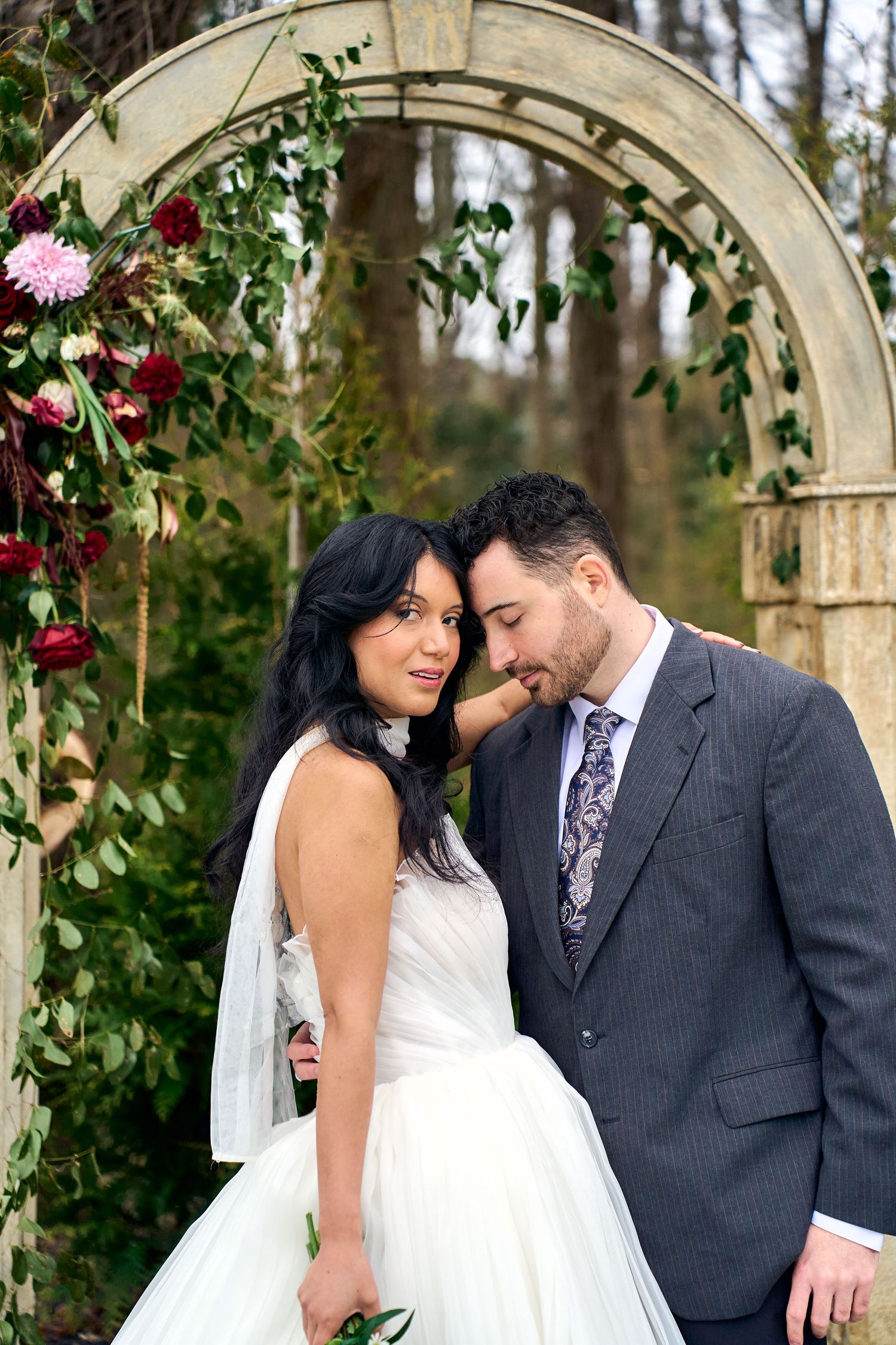 Romantic wedding couple poses under floral arch in outdoor garden setting with pink and red flowers.