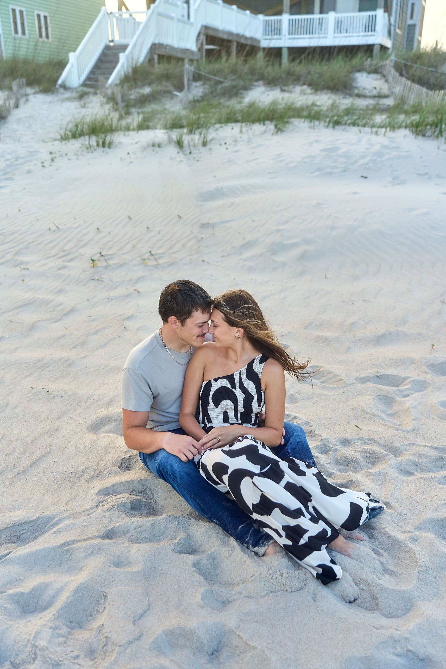 A couple sits together on a sandy beach near wood stairs and a beach house on a summer evening.