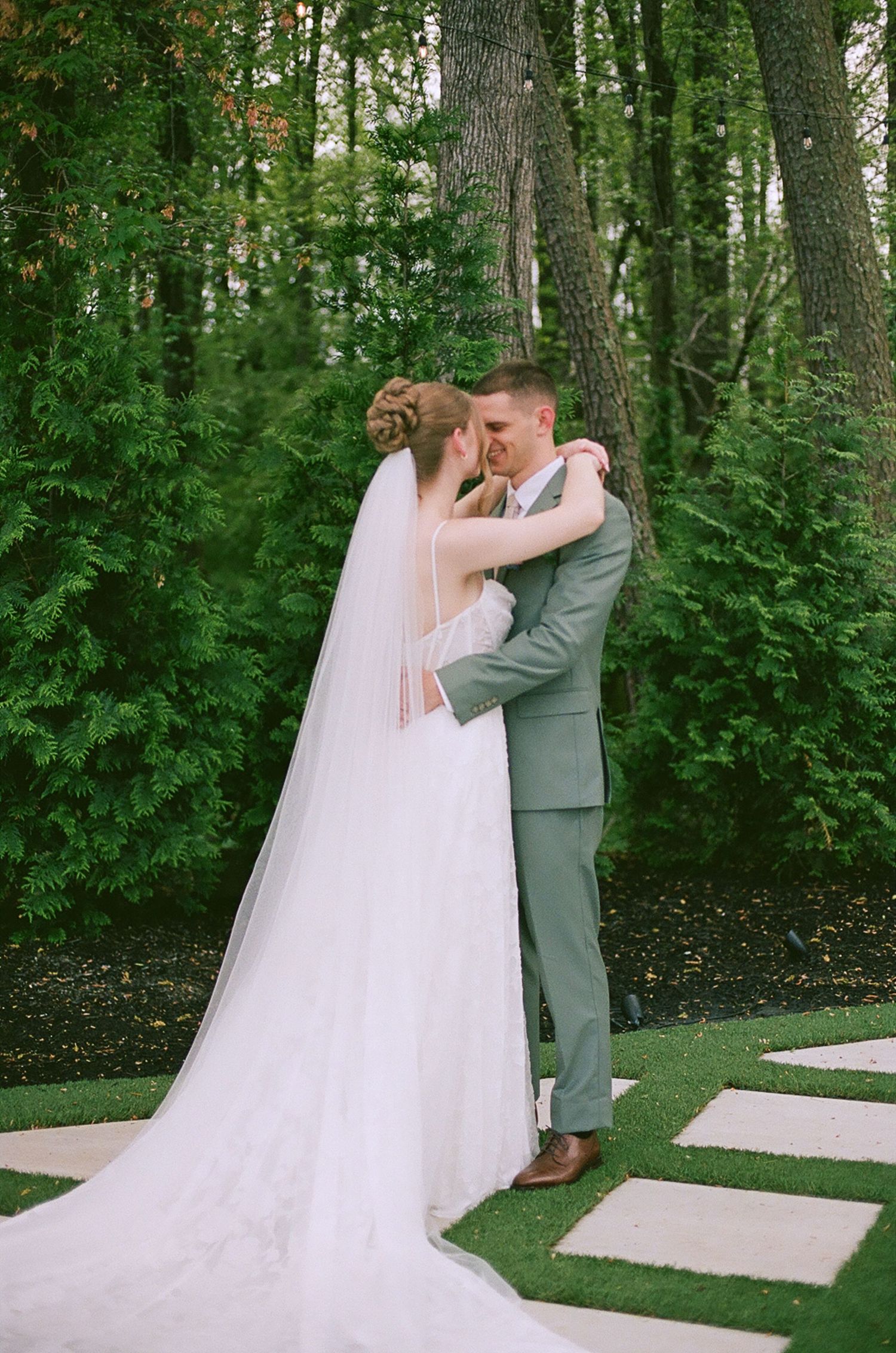 Bride in long veil and groom in sage suit embrace tenderly in lush green forest.