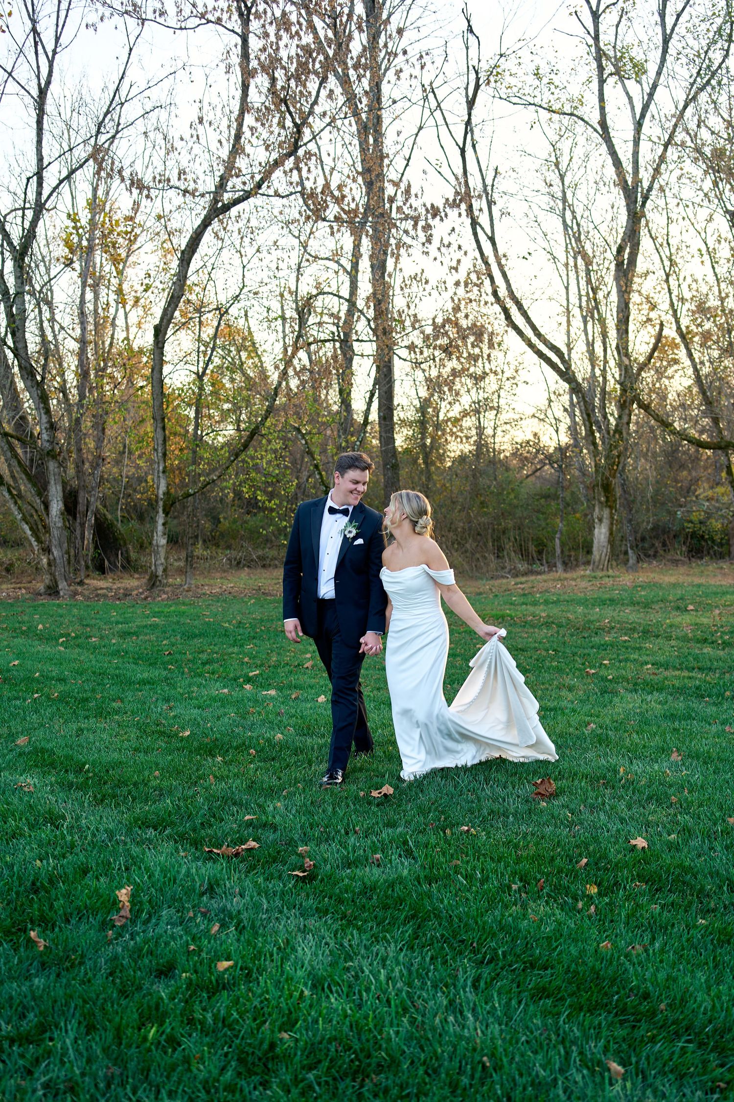 Bride and groom walk hand-in-hand across lush green lawn at golden hour sunset. North Carolina Wedding Photographer.