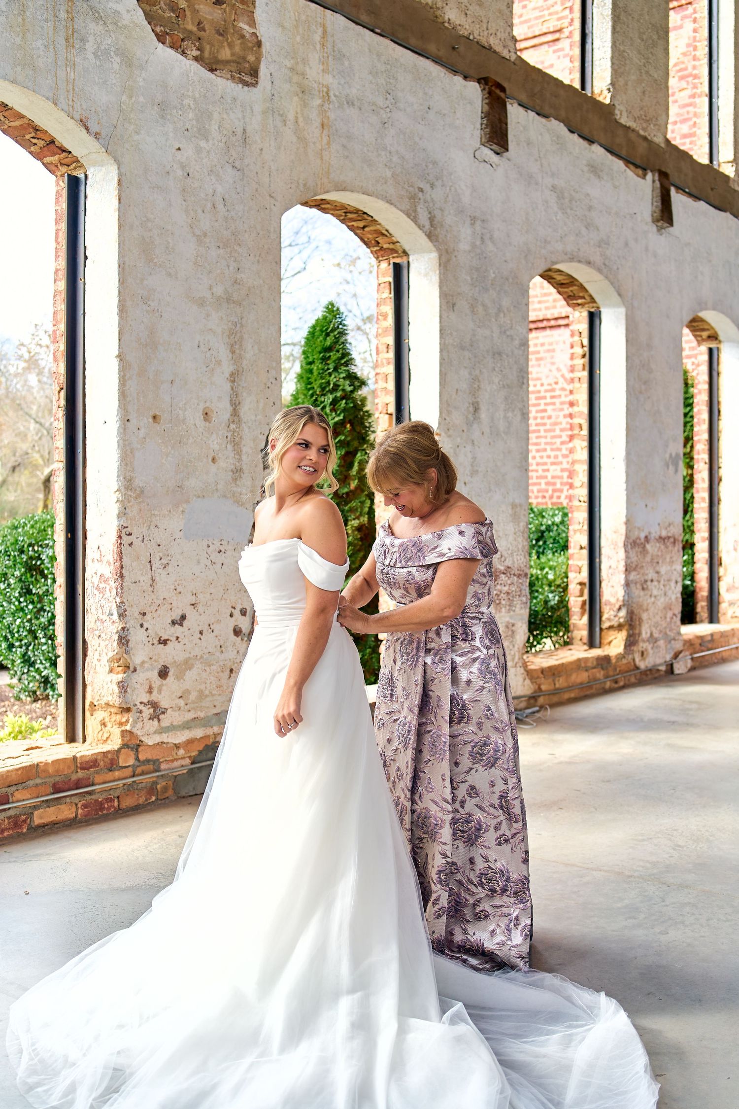 Two women stand in an elegant covered walkway with arched openings, one wearing a flowing white wedding gown with a long train.
