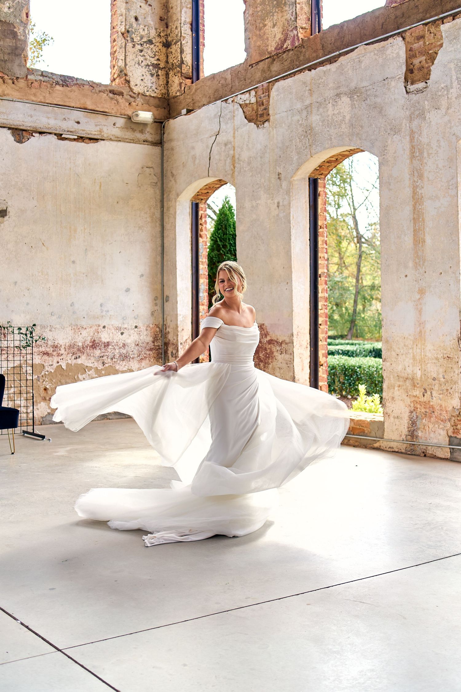 A flowing white wedding dress twirls in motion inside a rustic venue with arched windows and weathered walls.