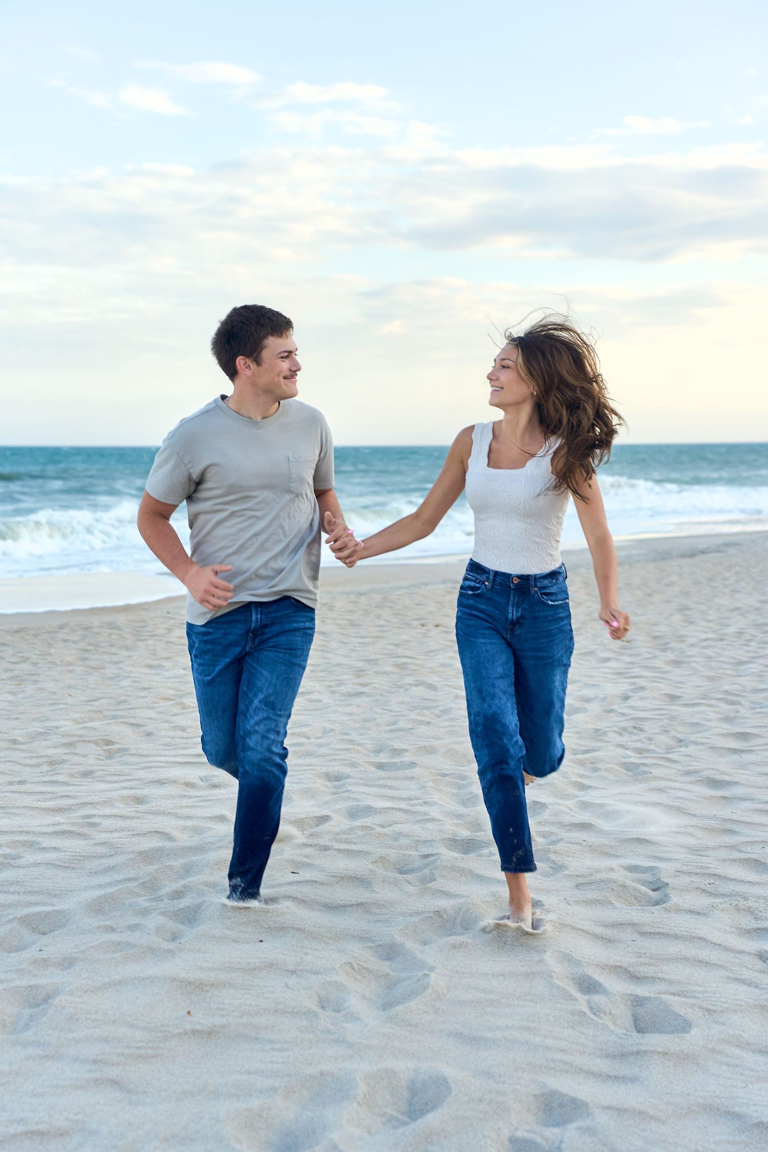 Two people in denim jeans run together playfully along a beach with ocean and sky in the background during sunset.