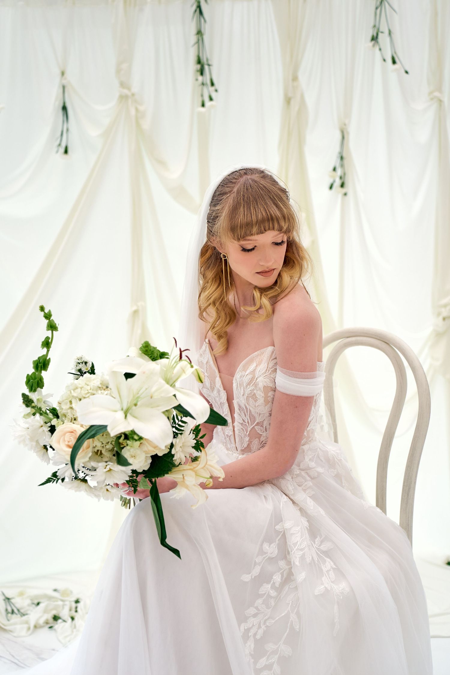 Elegant wedding dress with delicate lace details and off-shoulder sleeves shown in romantic setting with white floral bouquet.