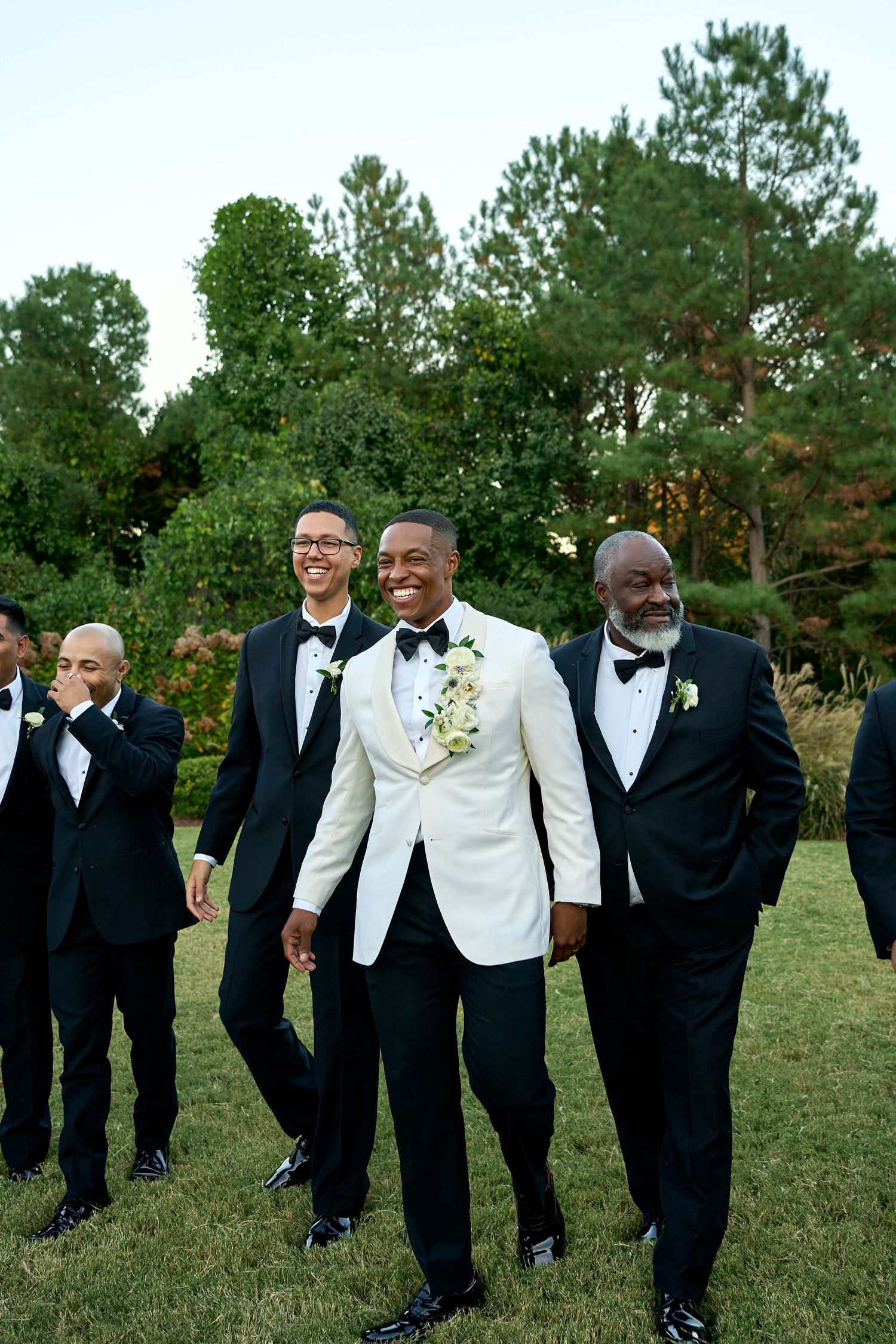 Groom in white tuxedo jacket smiles and walks with groomsmen in black tuxedos across a garden lawn.