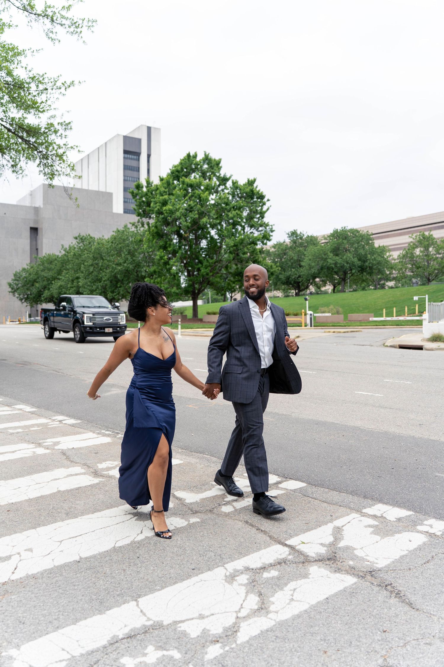 A stylish couple in formal wear holds hands while crossing a city street crosswalk together.