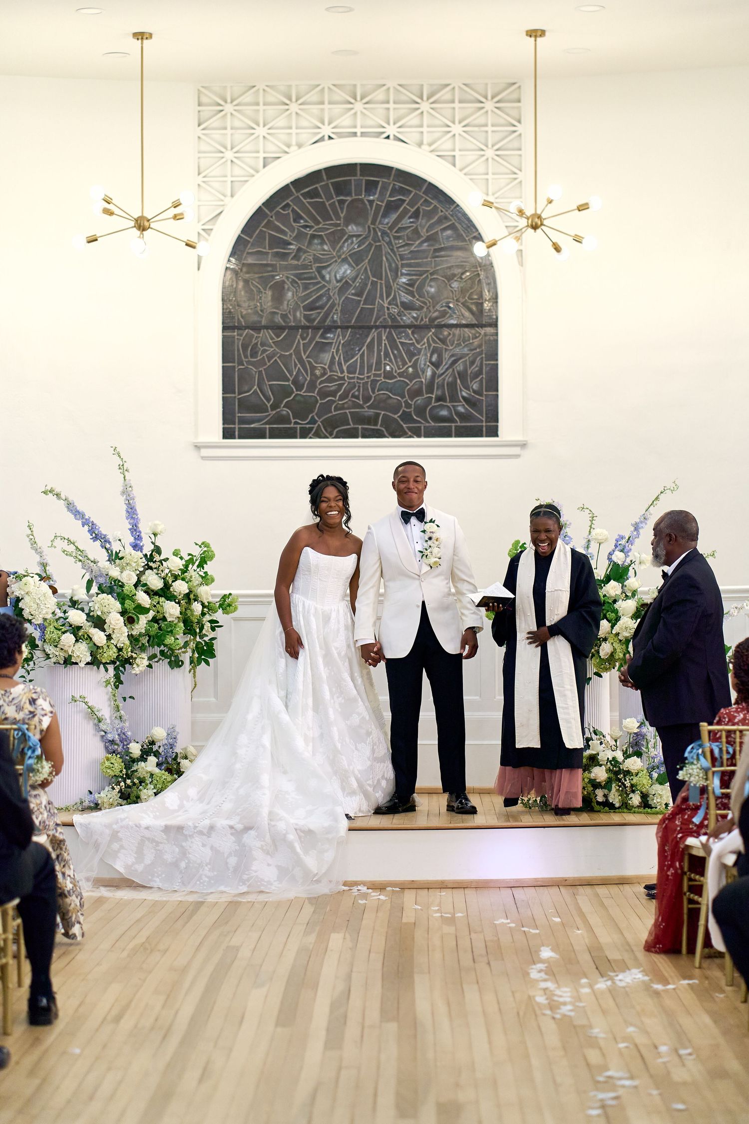 Bride and groom hold hands at altar during wedding ceremony in elegant white chapel with floral arrangements.