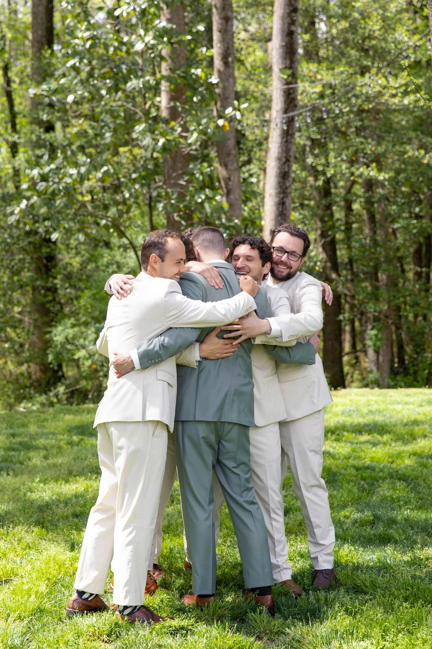 Groom in sage green suit shares a group hug with groomsmen in cream suits on green lawn.