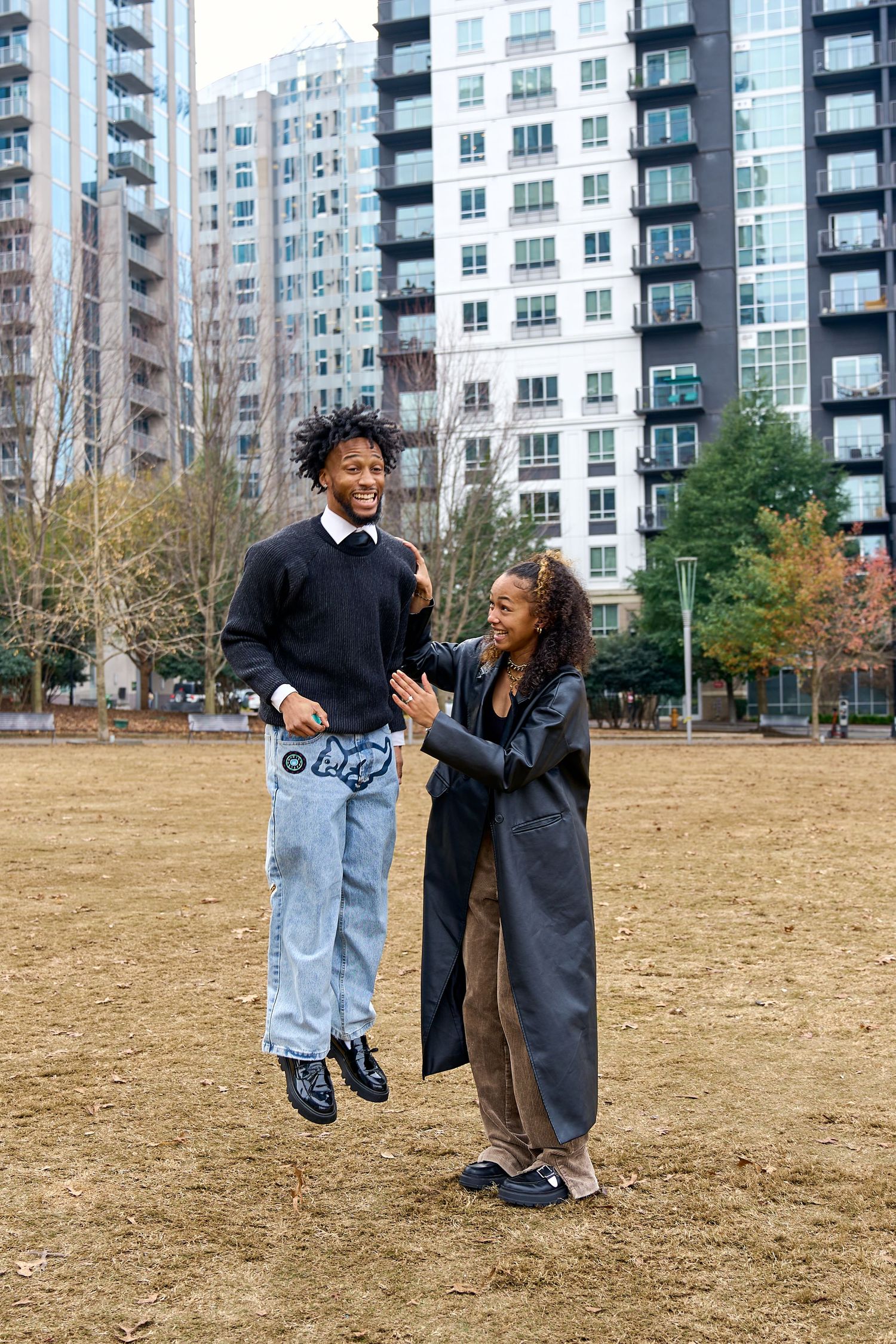 Two friends laugh and jump in an urban park with modern apartment buildings in the background. North Carolina Wedding Photographer.