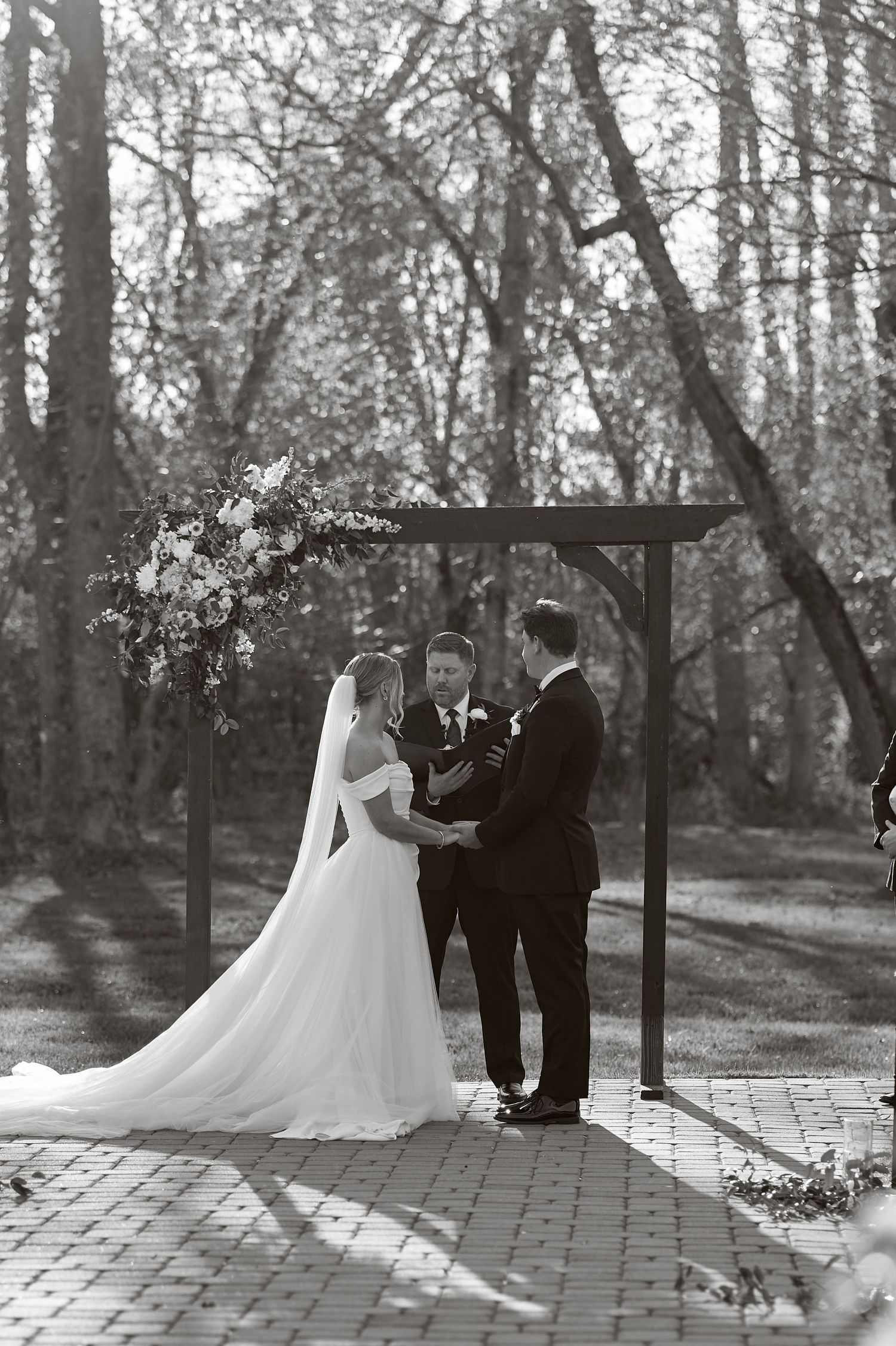 A wedding ceremony takes place under a wooden arch in a serene wooded setting, captured in black and white photography.