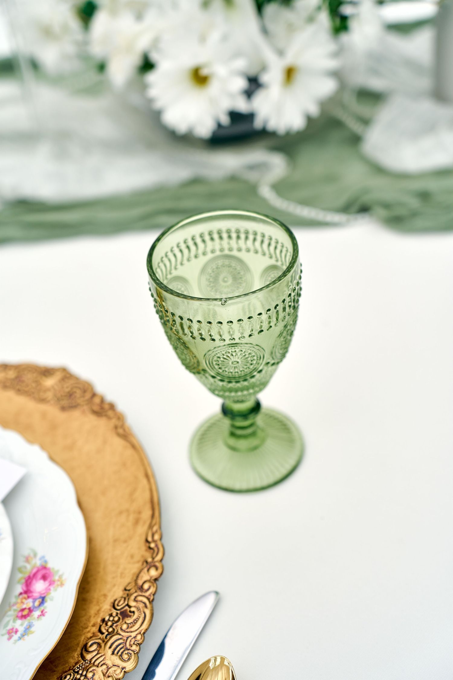 A vintage-style green glass goblet sits beside a gold-rimmed plate with white daisies in the background.