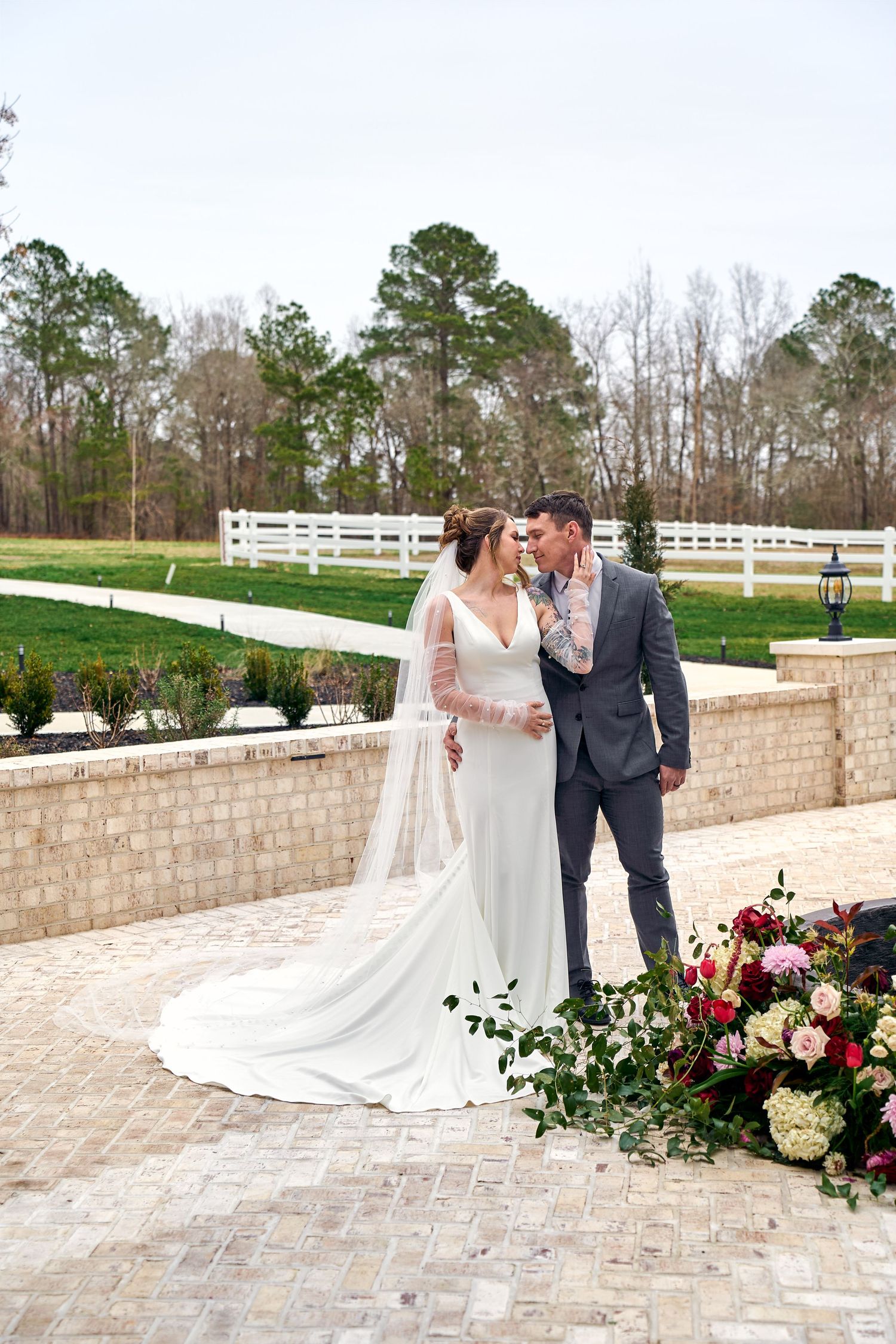 White wedding floral display with a couple on a brick paved patio with white fencing and green trees in background.