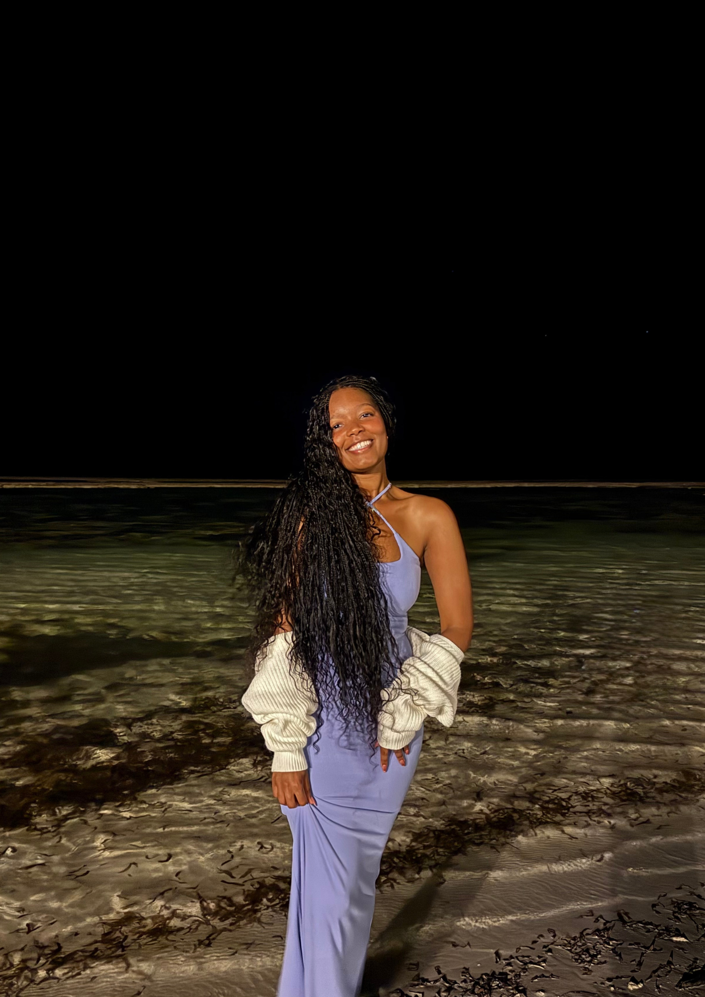 Smiling woman in lavender dress with long braids standing at the ocean's edge at night.