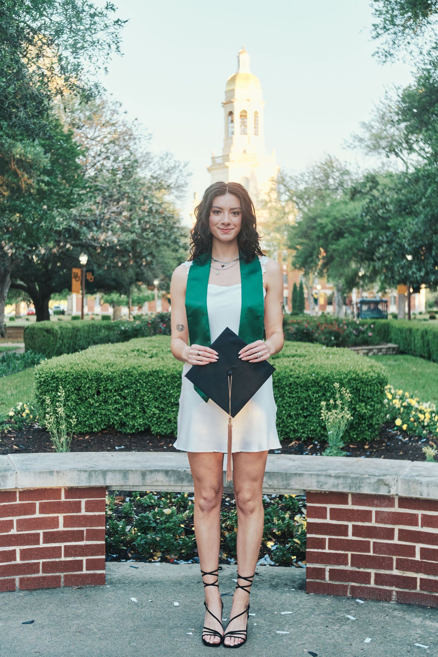 Young woman in white dress holding graduation cap with green stole near Baylor chapel.