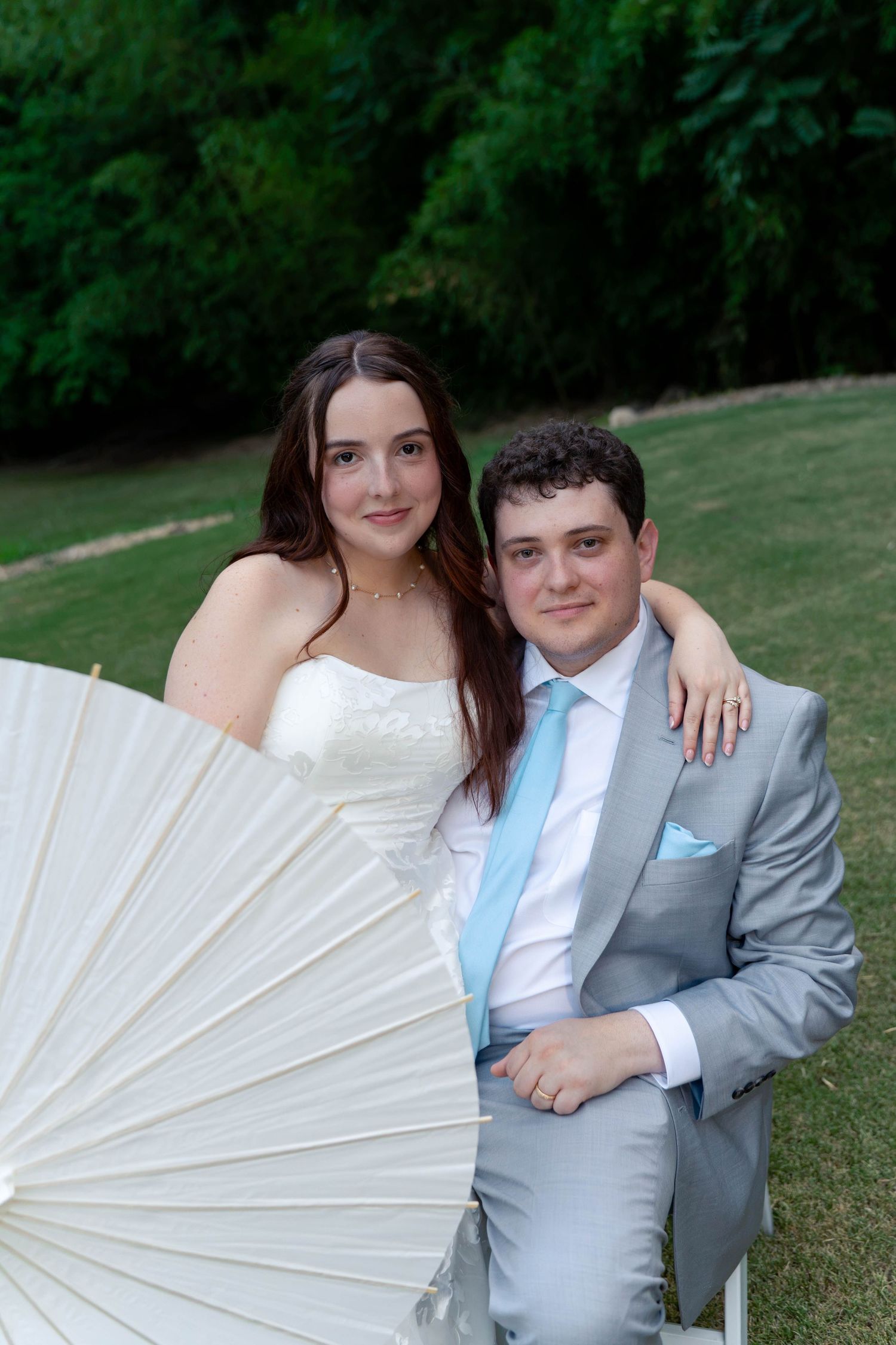 Bride and groom pose with white parasol umbrella while seated outdoors at their garden wedding ceremony.