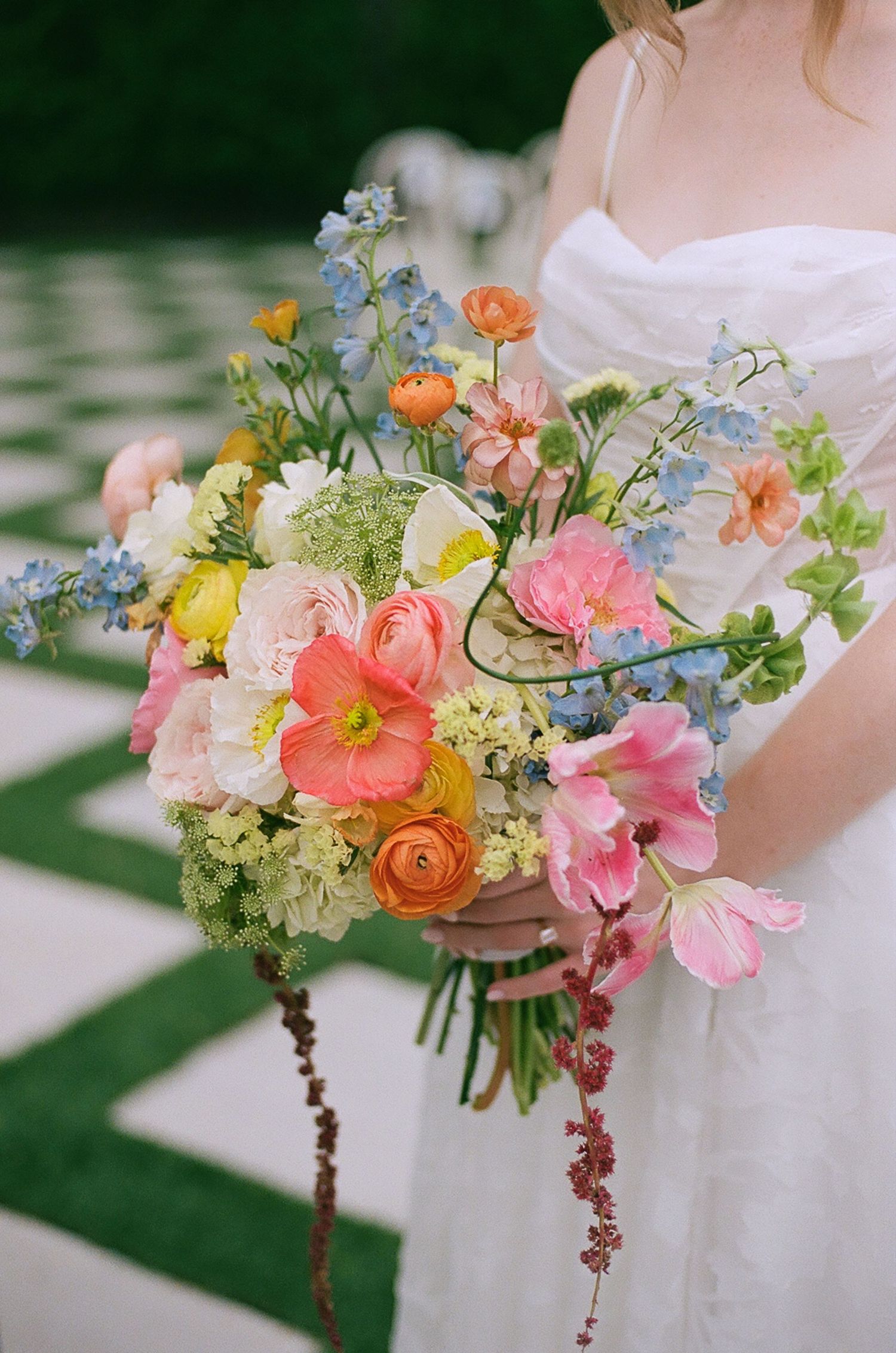 Bride holds vibrant wildflower bouquet with pink, blue, orange, and yellow blooms outdoors.