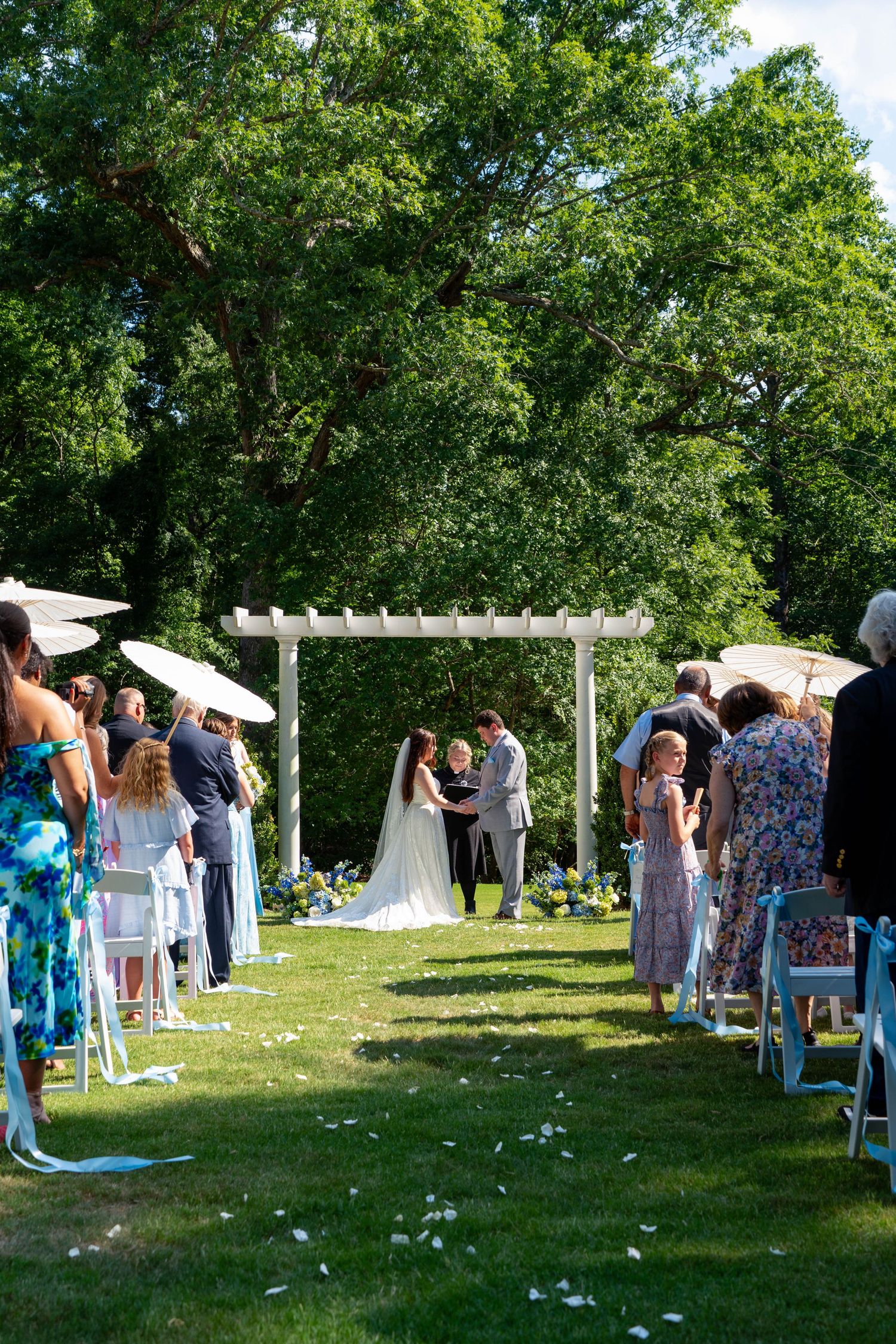 Outdoor wedding ceremony under white pergola arch with guests seated on white chairs on lawn.