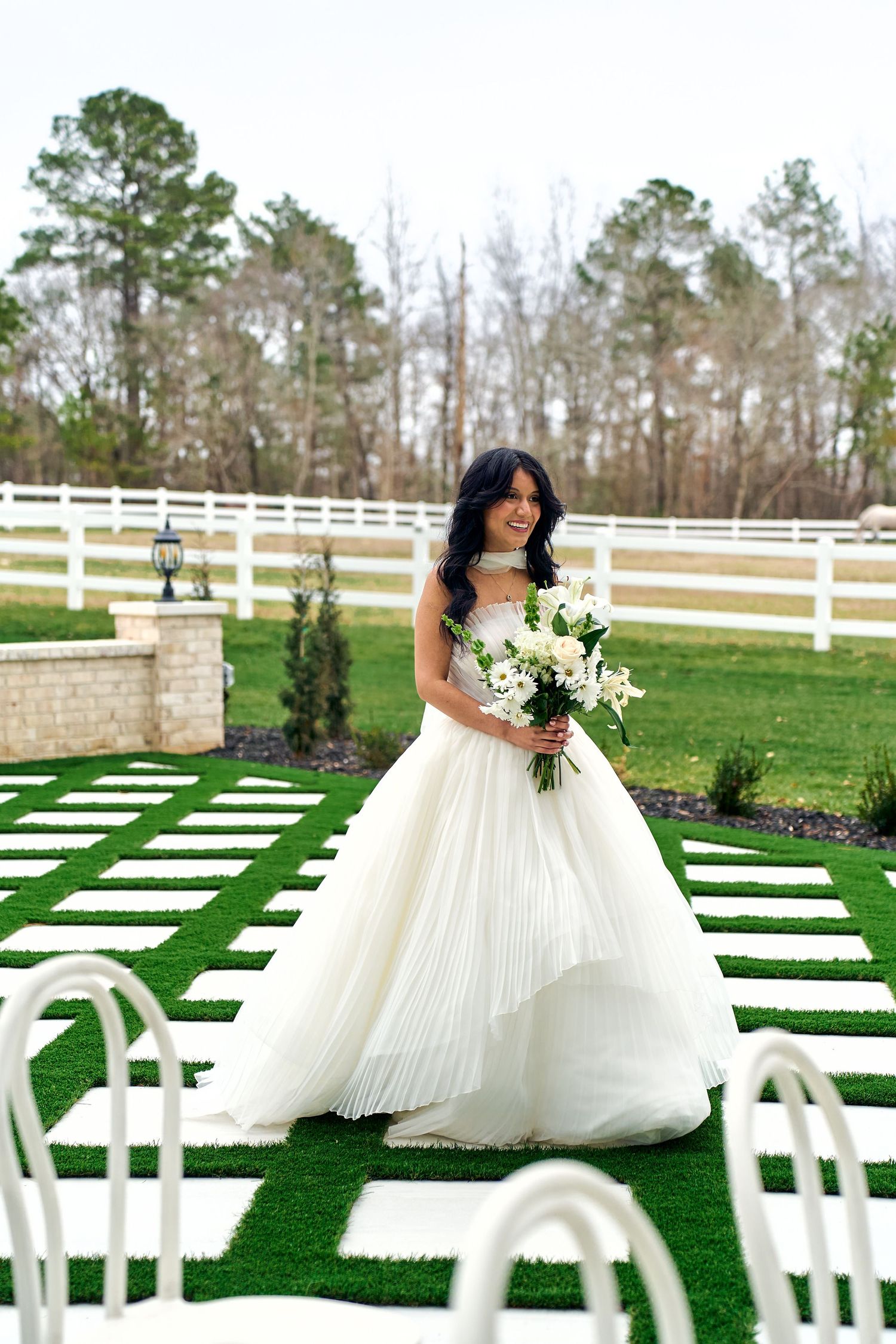 Beautiful outdoor wedding ceremony setup with white chairs on green grass and white fence with tall pine trees in background.