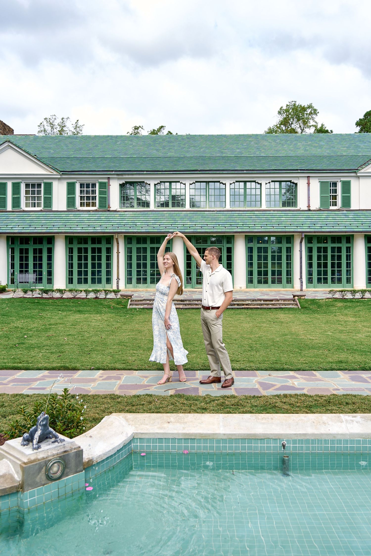 Couple dancing by a pool in front of elegant green-roofed estate on sunny day.