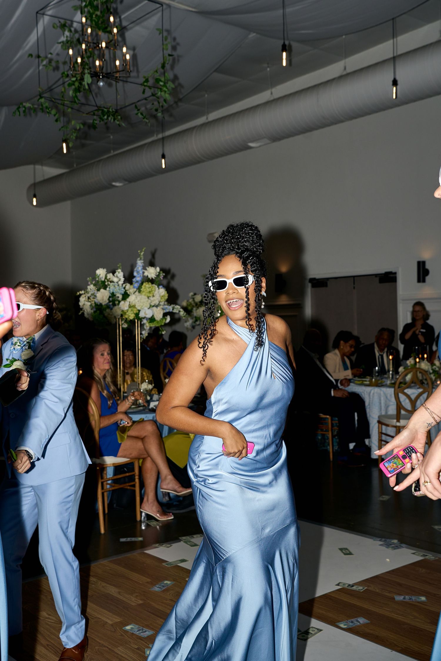 Wedding guest in blue satin gown and white sunglasses dances at elegant indoor wedding reception.