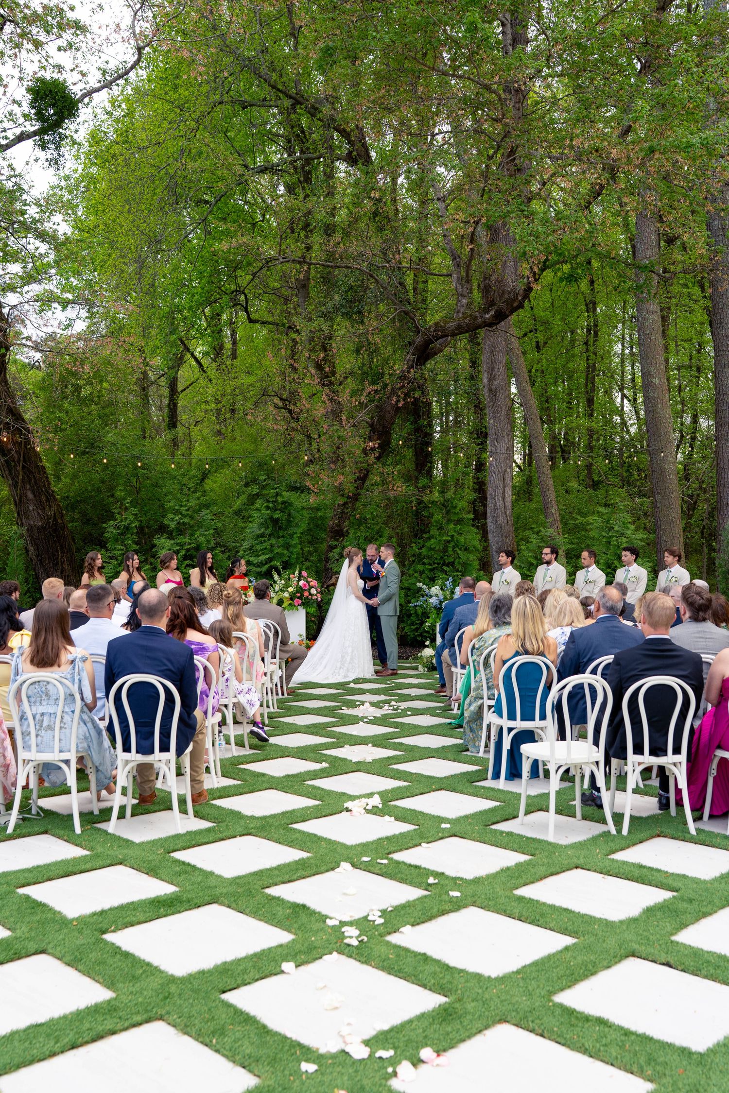 Guests seated on white chairs watch bride and groom exchange vows at lush outdoor garden wedding.