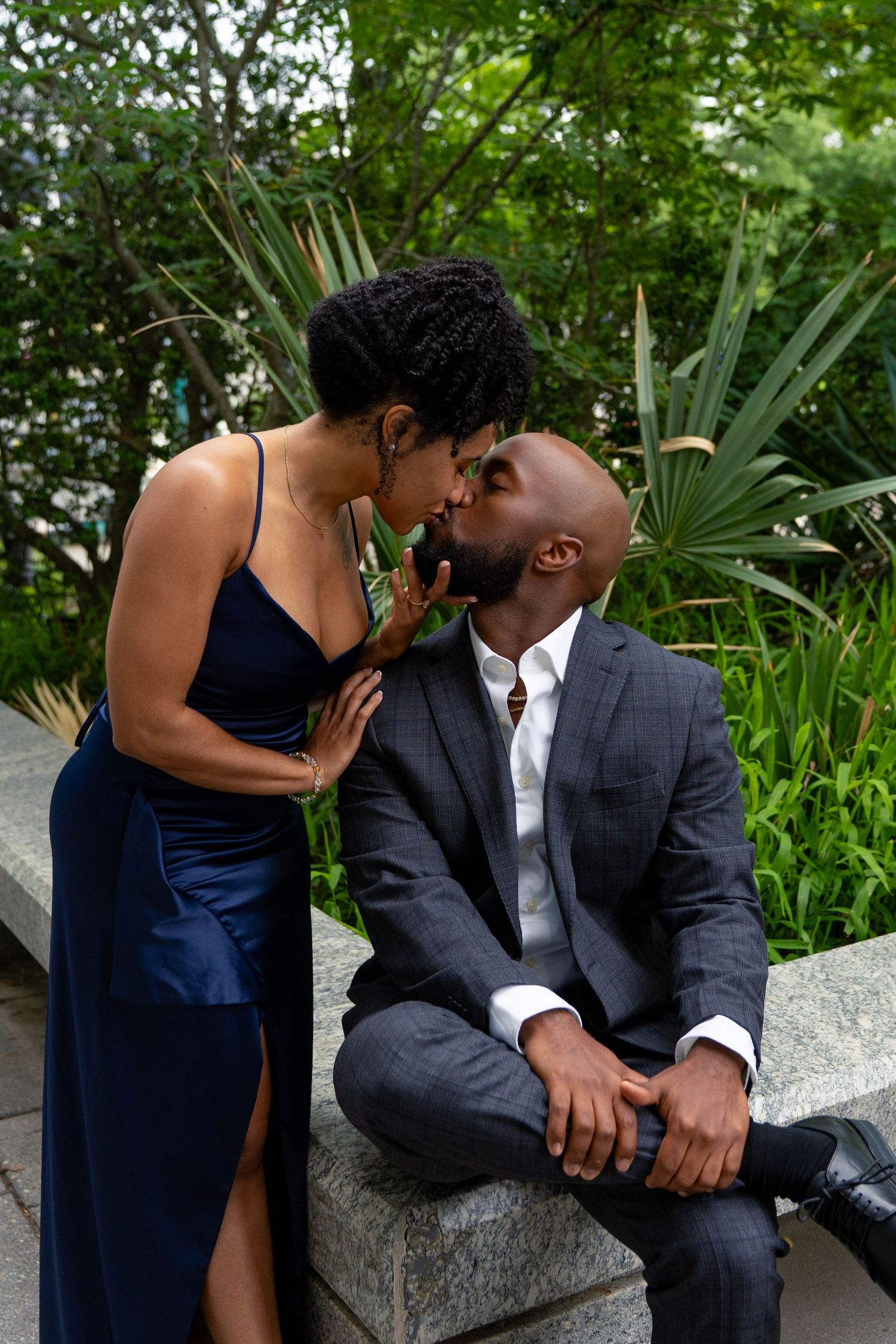 Romantic Black couple sharing a kiss on a stone bench surrounded by lush green garden foliage.