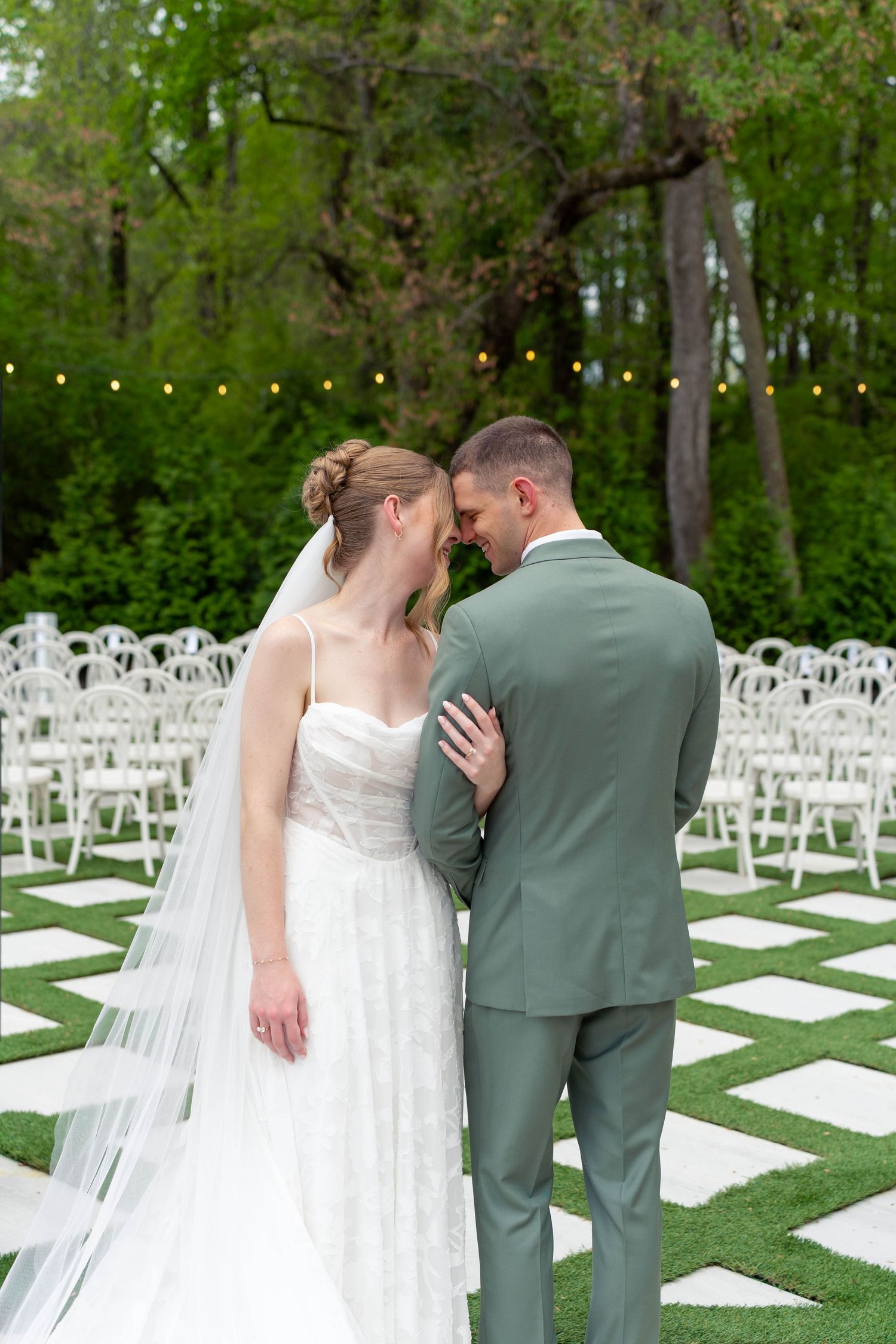 Bride and groom share an intimate moment touching foreheads at an outdoor garden wedding venue with string lights.