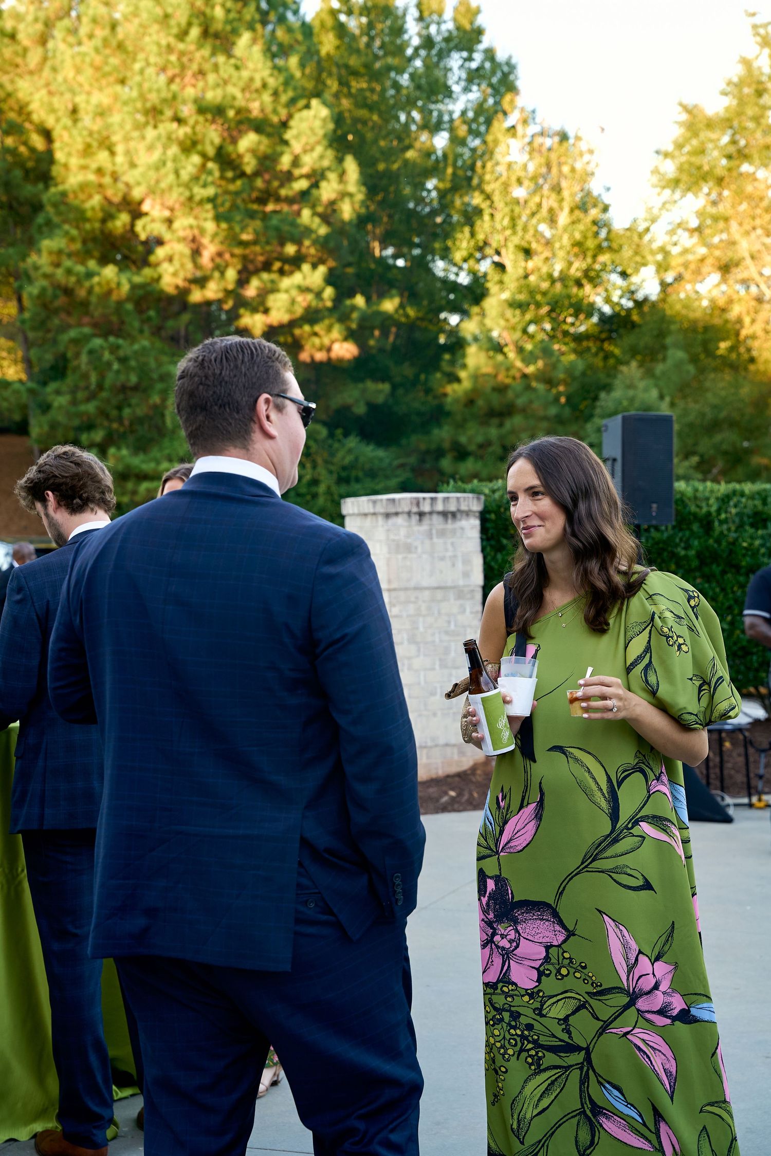 Wedding guest in green floral dress converses with man in navy suit during outdoor cocktail hour.