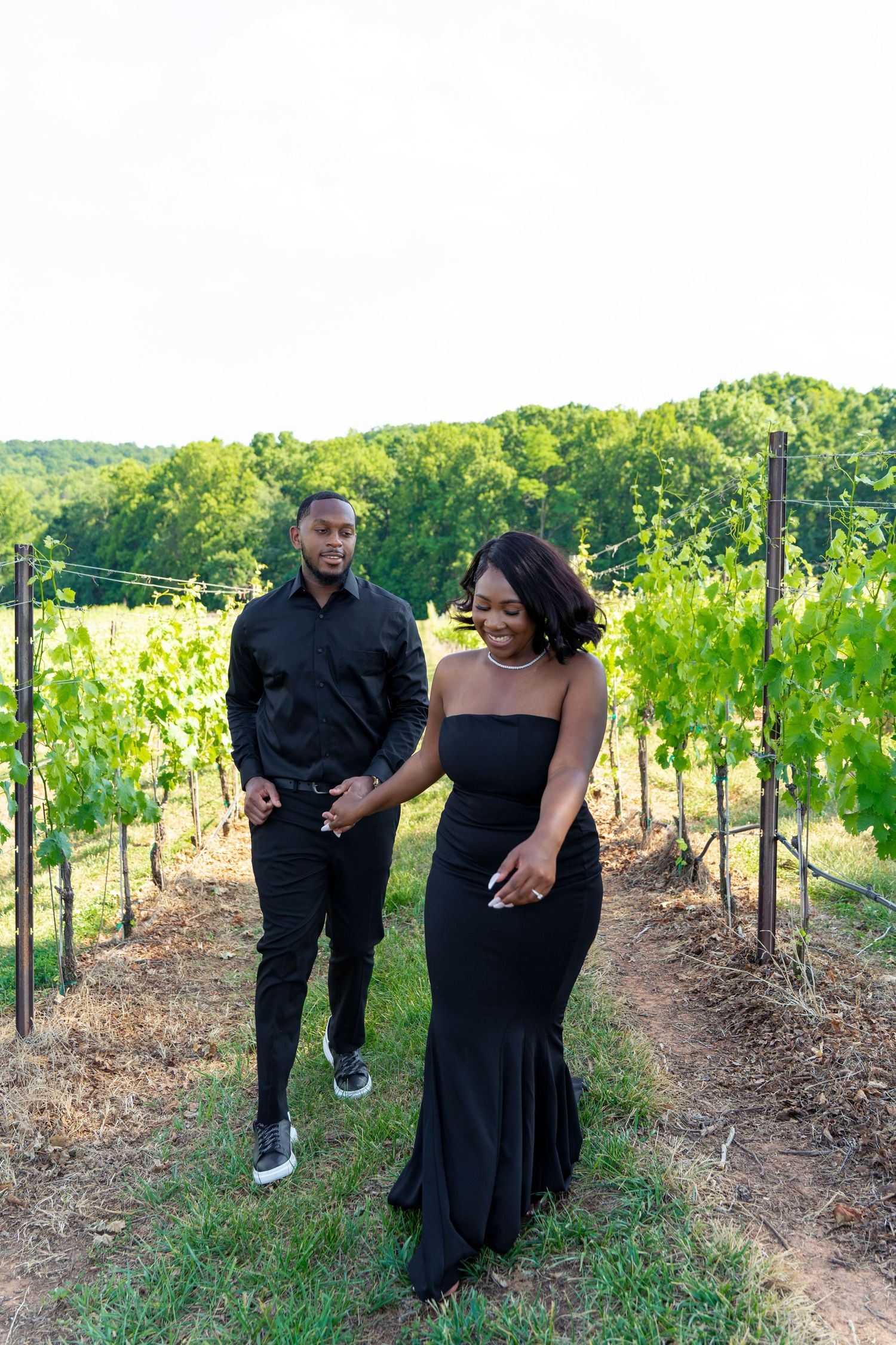Elegant couple in black attire walk hand in hand through lush green vineyard rows.