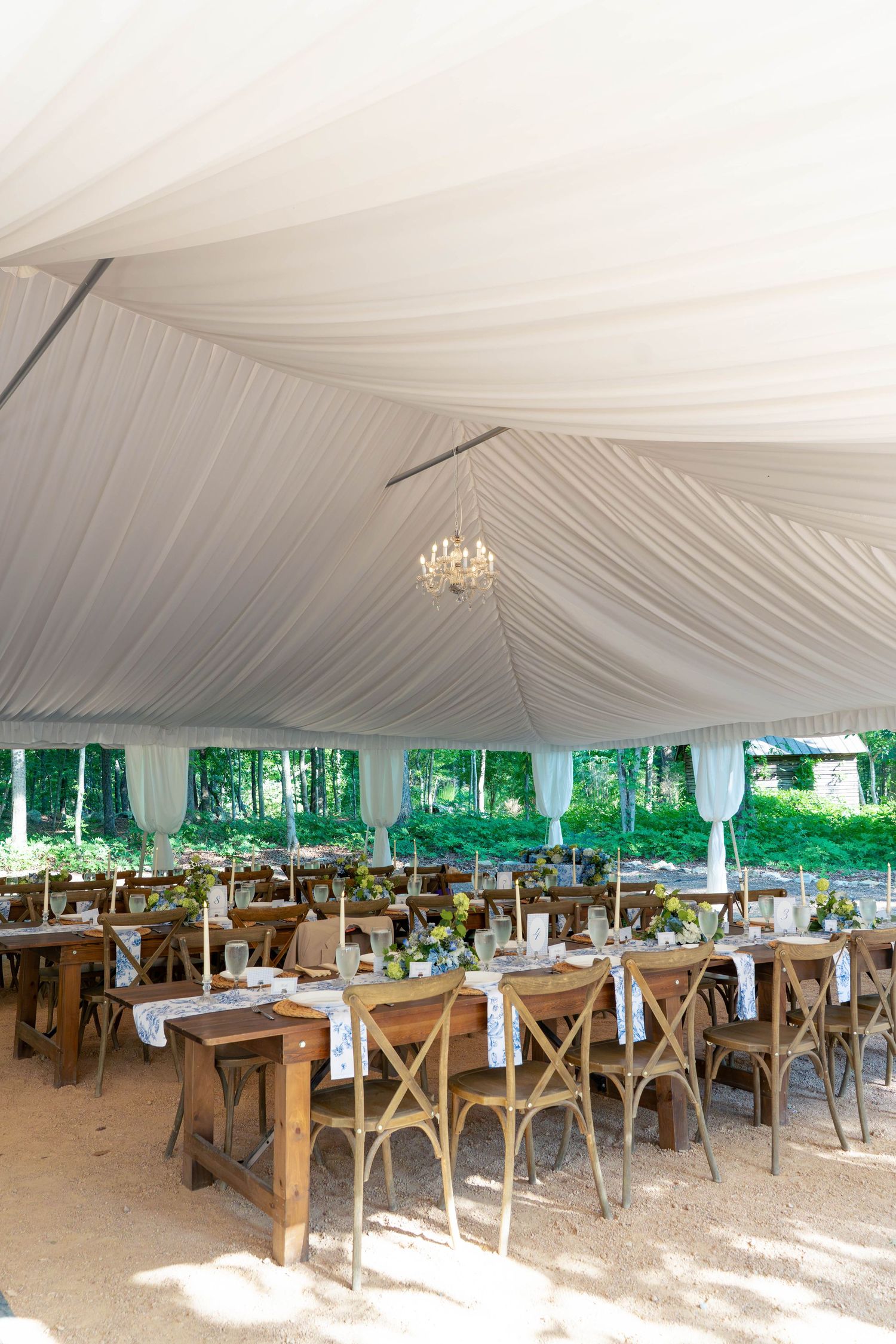 Elegant wedding reception tent with draped white ceiling, chandelier, and rustic wooden tables.
