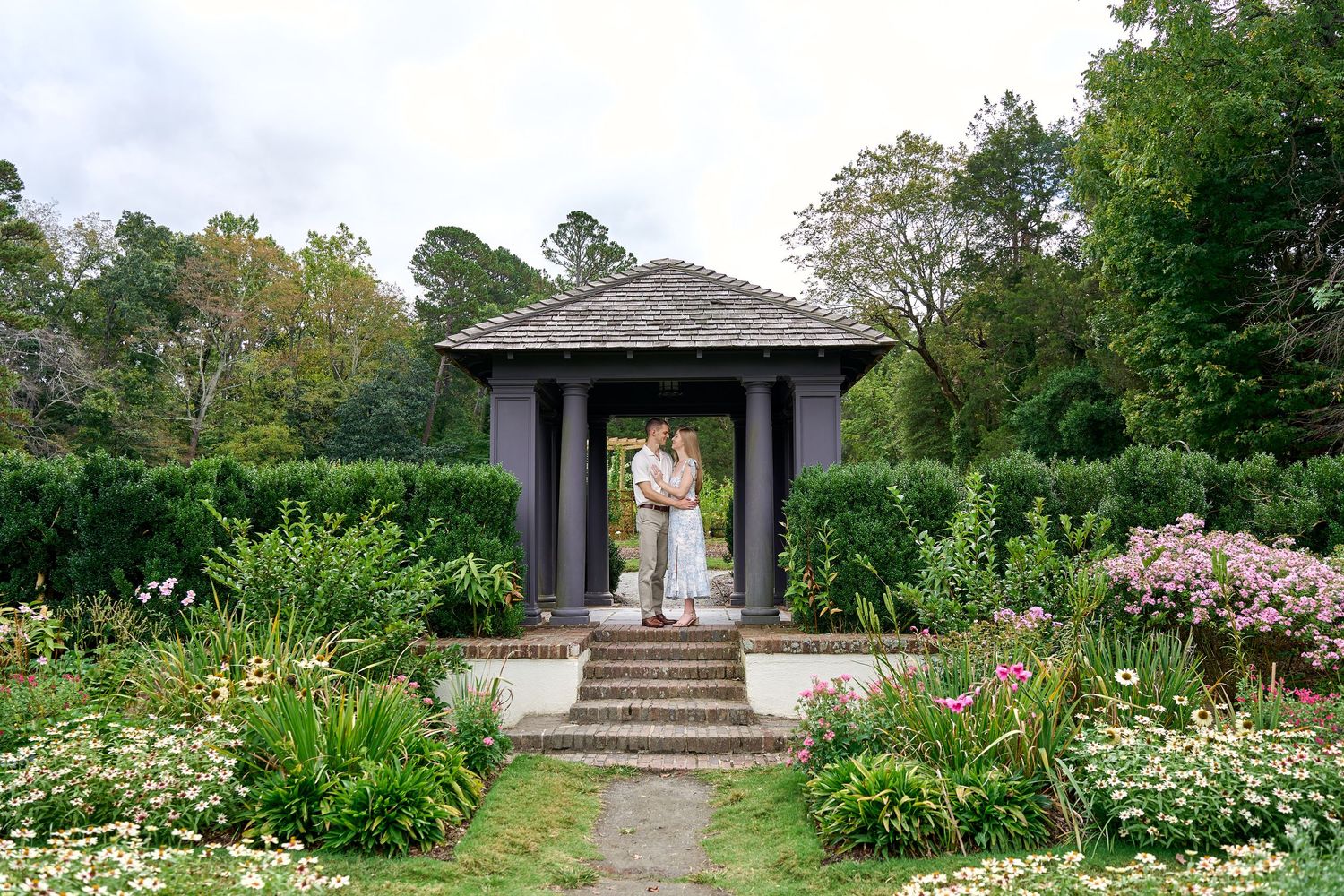 A dark wooden gazebo surrounded by lush gardens and flowering plants in a serene outdoor setting.